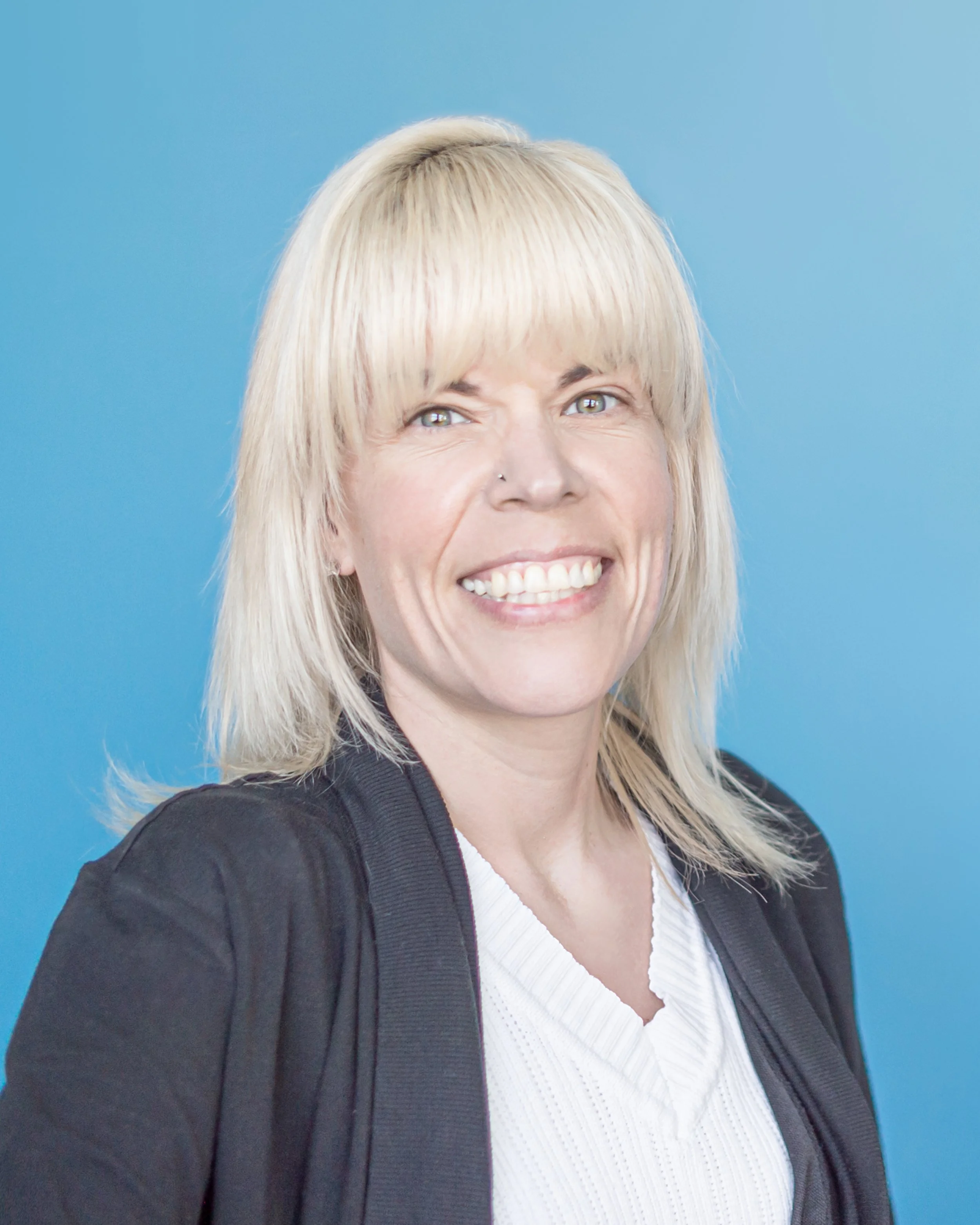 Close-up of a smiling woman with blonde hair and light green eyes, wearing a black jacket over a white sweater, standing against a blue background.