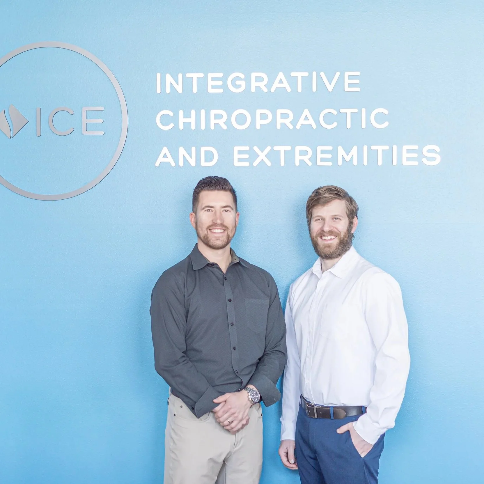 Two men standing together in front of a blue background. They are smiling and dressed in business casual attire, with one in a dark shirt and the other in a white shirt. Text on the background reads 'Integrative Chiropractic and Extremities'.