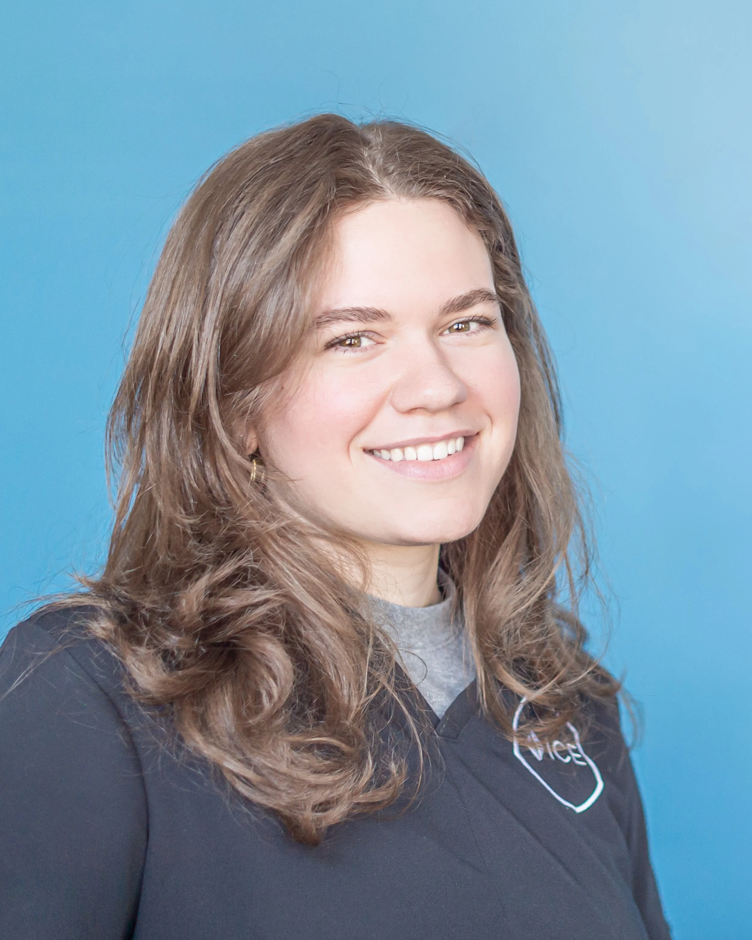 Close-up of a young woman with long, wavy brown hair smiling, wearing a black hoodie with a white outline of a smiley face and the word 'NICE' on it, against a bright blue background.