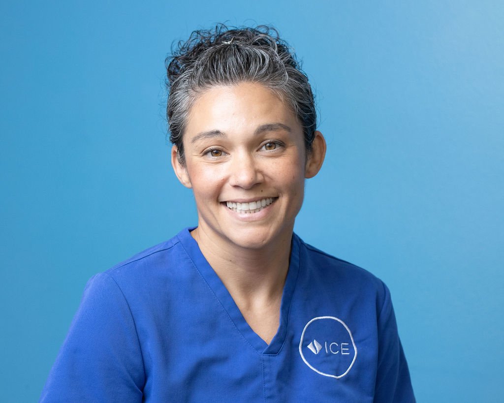 A woman with short curly gray hair smiling at the camera, wearing blue medical scrubs with an ICE logo embroidered on the chest, against a blue background.