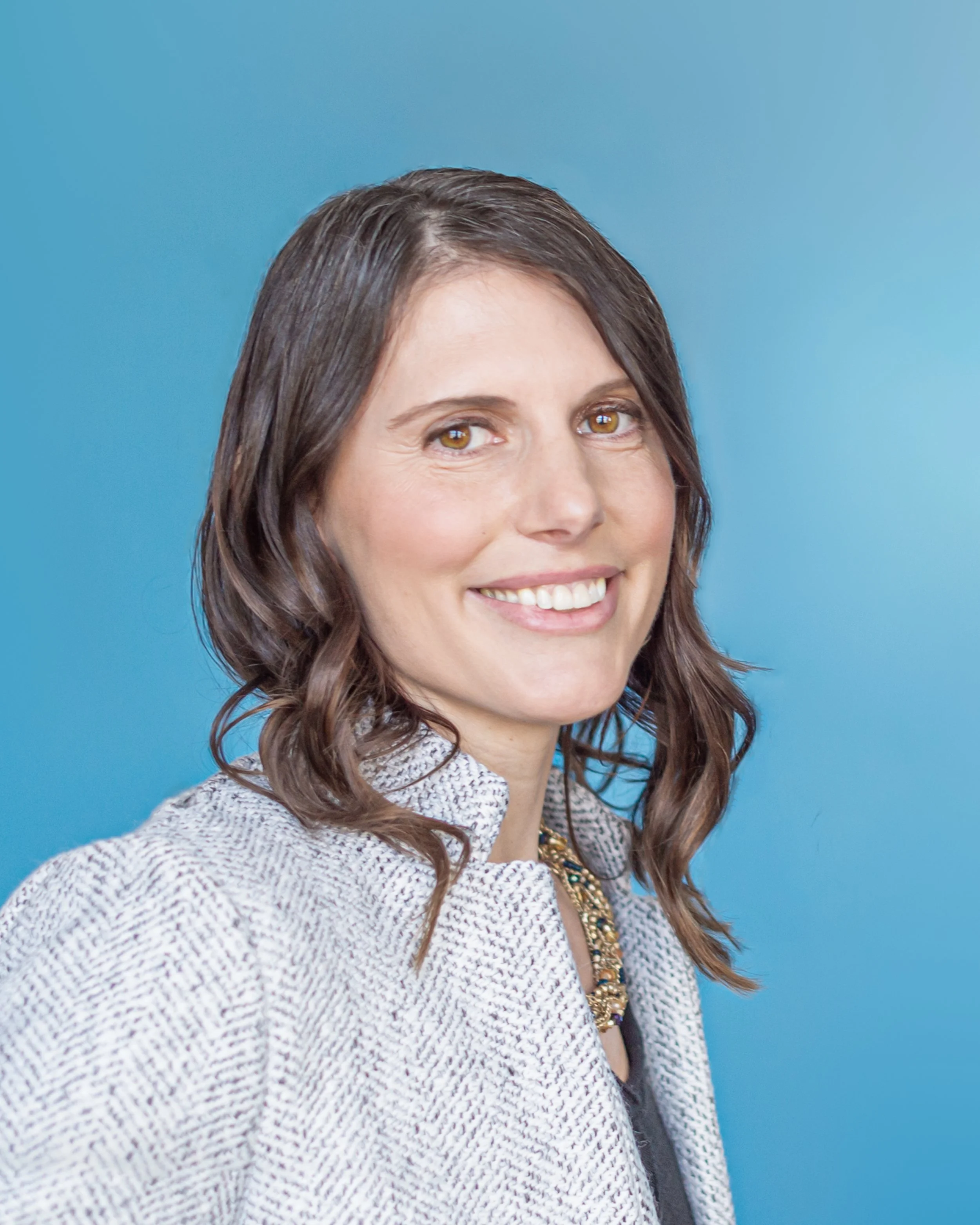 A smiling woman with wavy brown hair, hazel eyes, wearing a patterned blazer and a necklace, standing against a light blue background.