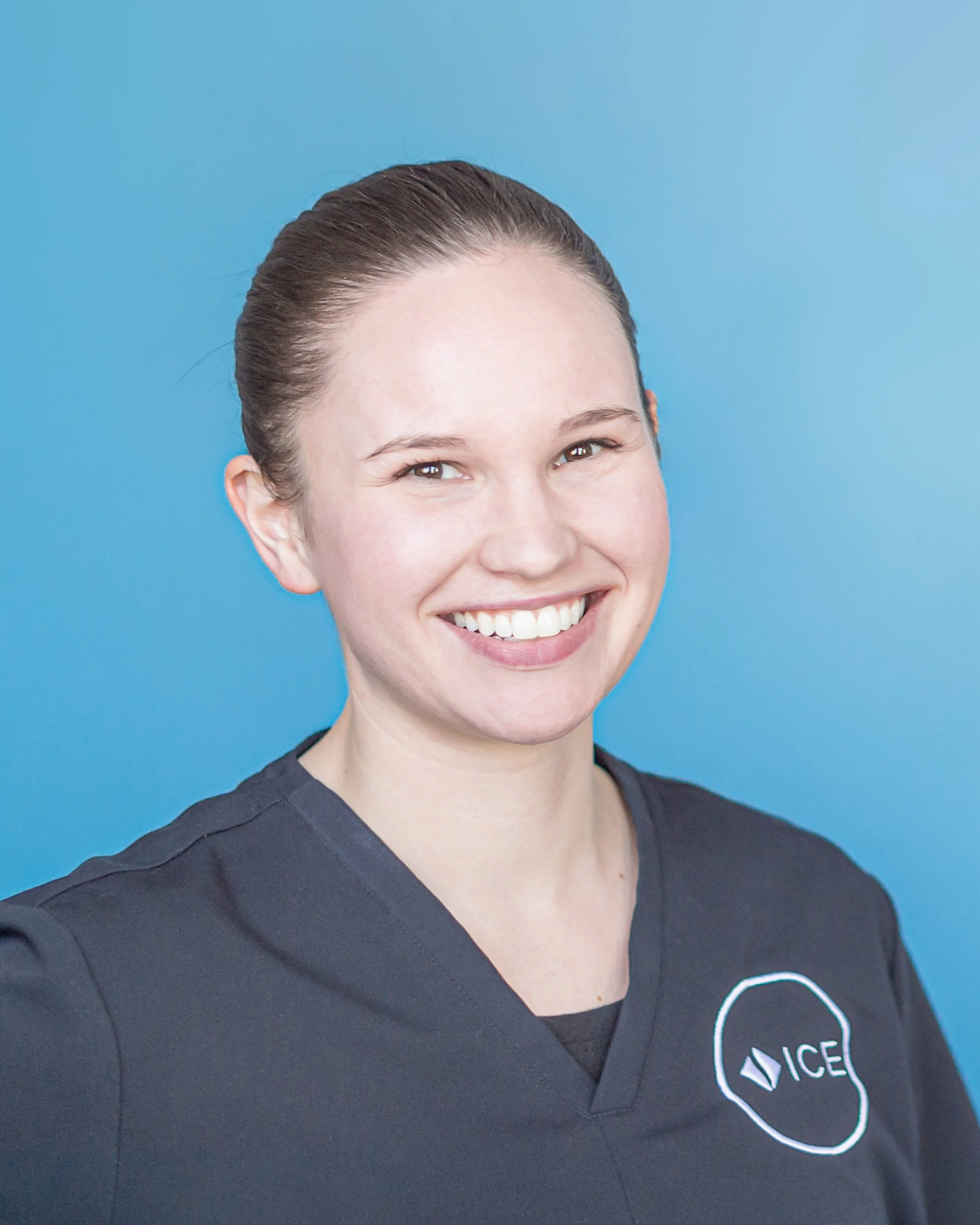 Close-up of a woman smiling, wearing a black t-shirt with a circular white logo that says 'ACE' and has a geometric diamond shape inside the circle against a blue background.