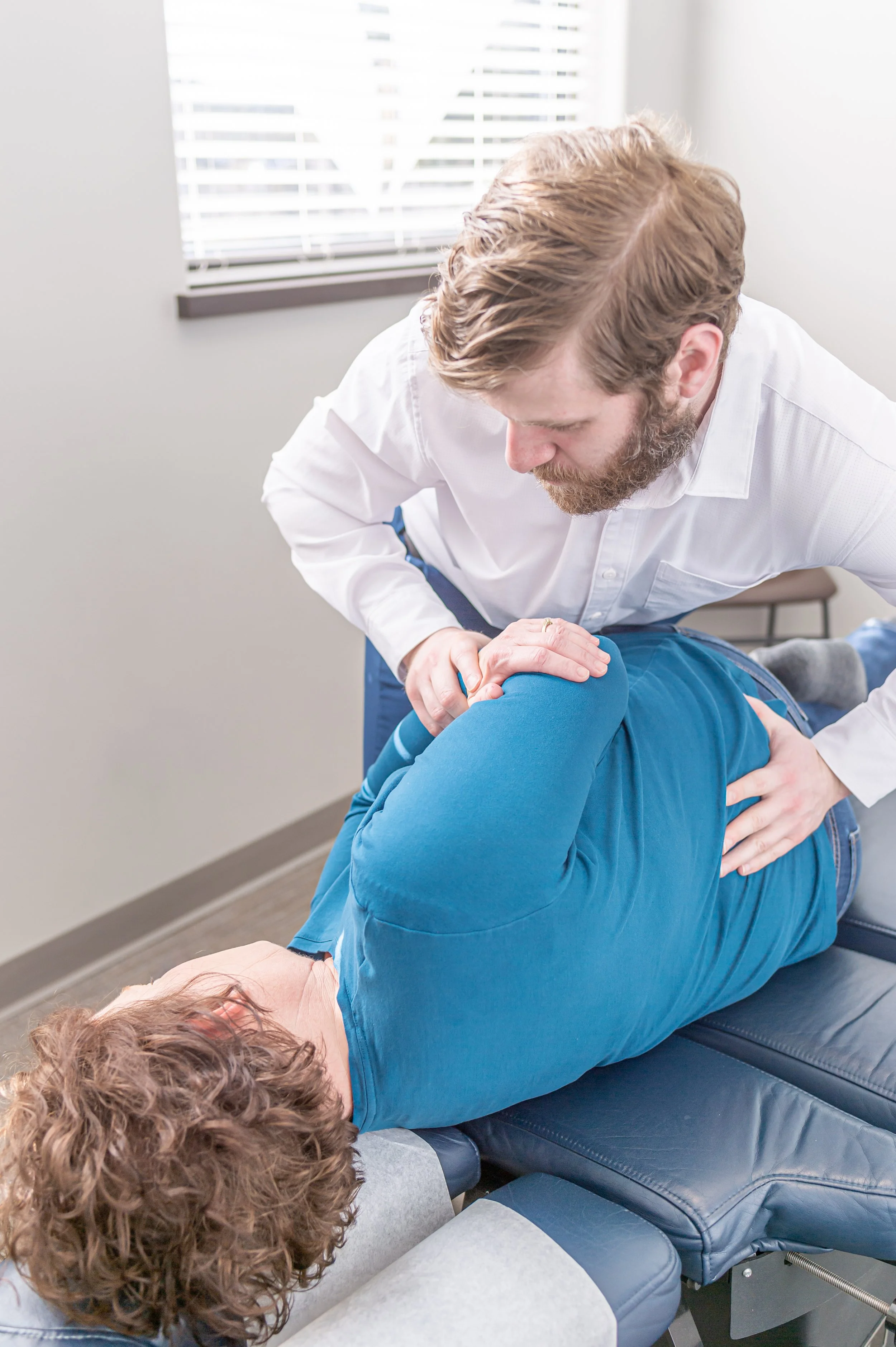 Dr Joe adjusts a patient's back as the patient lies face down on a treatment table in a medical office with a window and blinds in the background.