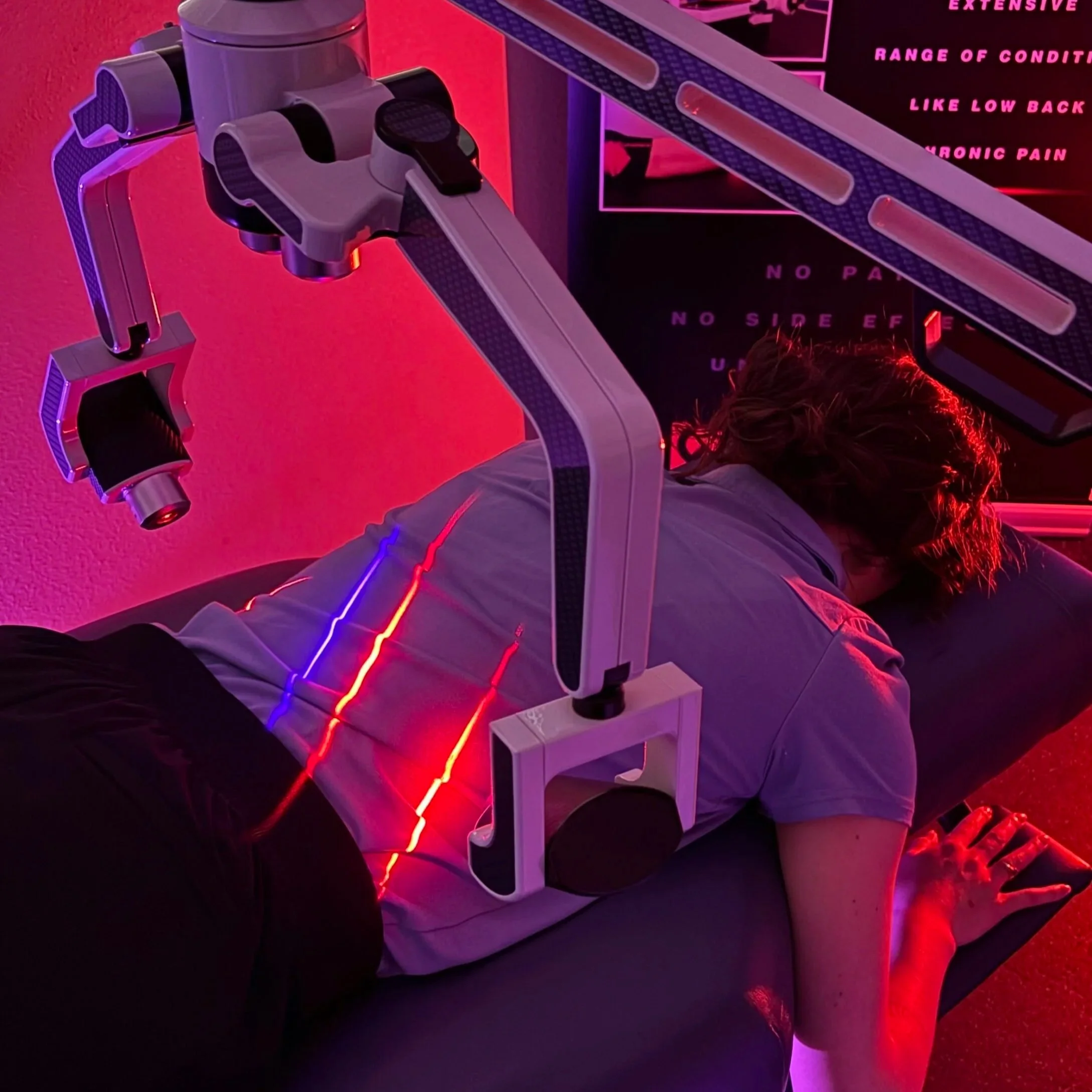 Close-up of a medical robotic arm with multiple joints and attachments, illuminated by colorful pink and purple lighting, with a person lying on a medical bed nearby.