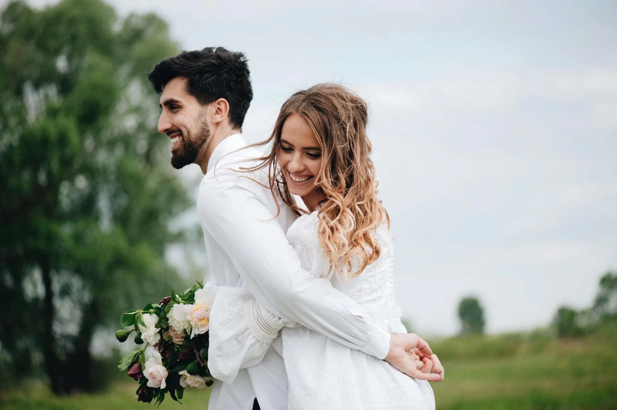 A smiling couple, a man with dark hair and beard and a woman with wavy blonde hair, embracing outdoors on a cloudy day, holding a flower bouquet.