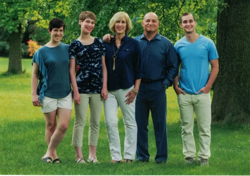 Family of five posing outdoors on a grassy field with trees in the background. Two women and three men, all smiling.