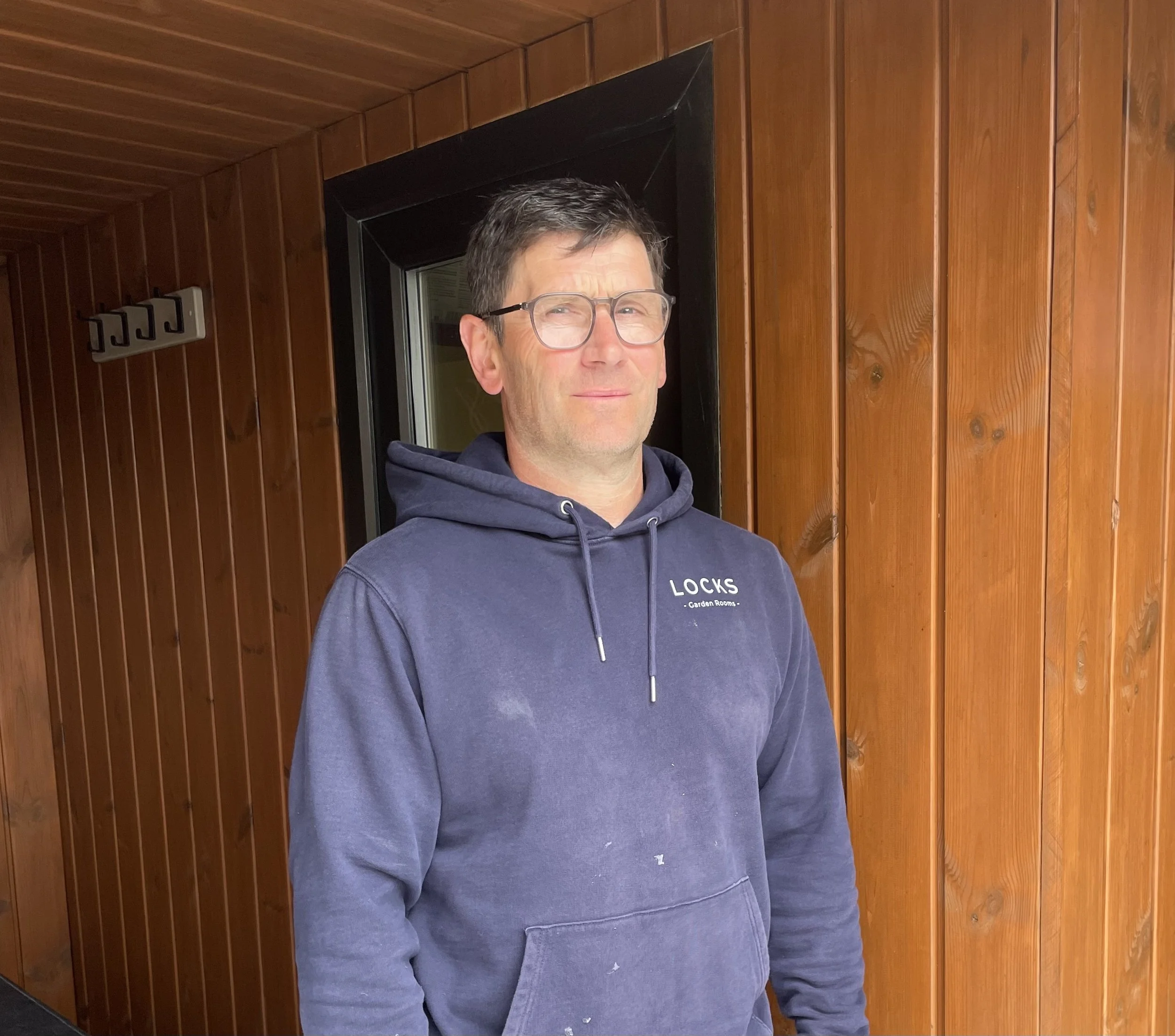A middle-aged man with glasses wearing a navy blue hoodie, standing in front of a wooden wall with a window behind him.