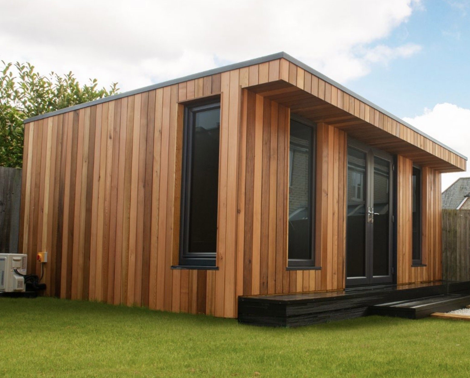 A modern tiny house with wooden vertical siding, large glass doors and windows, situated on a green lawn with a cloudy sky overhead.