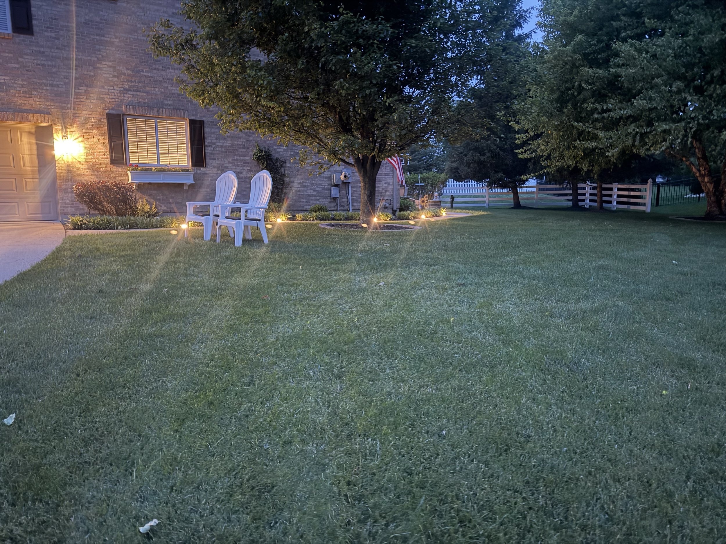 A backyard scene in the evening featuring a large green lawn, two white chairs near a tree, a brick house with a window and a light on, and a white fence in the background. There are decorative lights along the garden bed and an American flag hanging