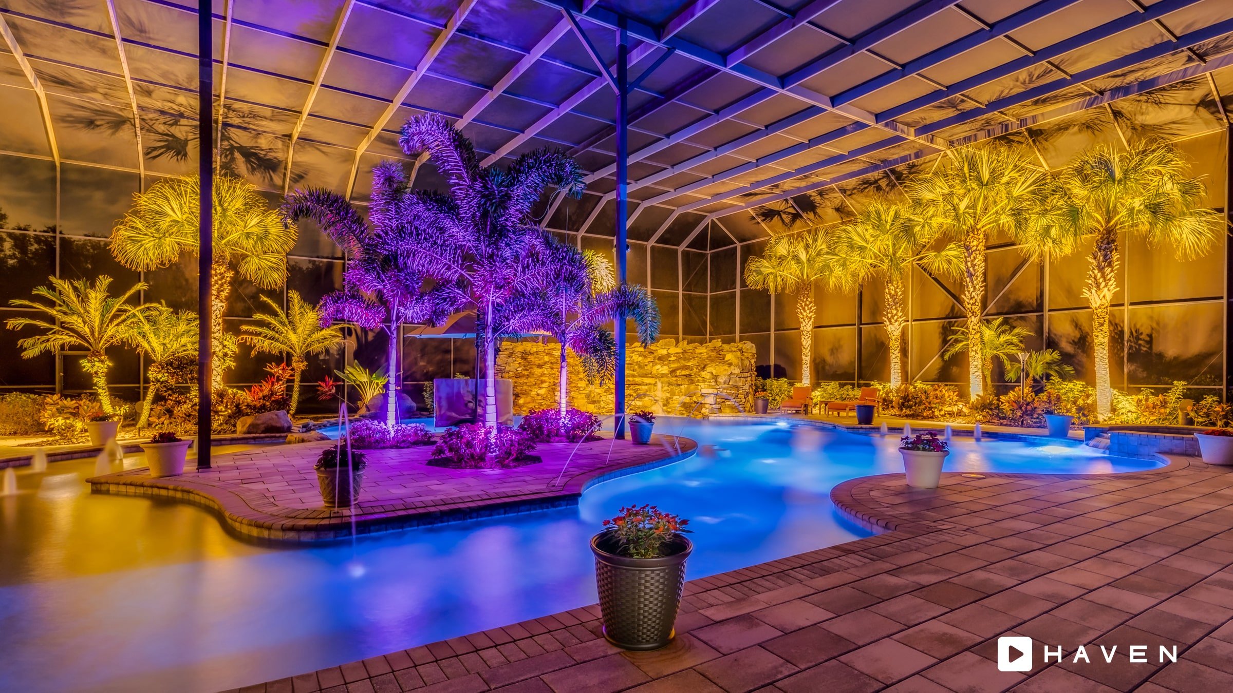 An enclosed backyard pool area illuminated with colorful lights, surrounded by palm trees and potted plants, with a fountain feature and a brick patio.