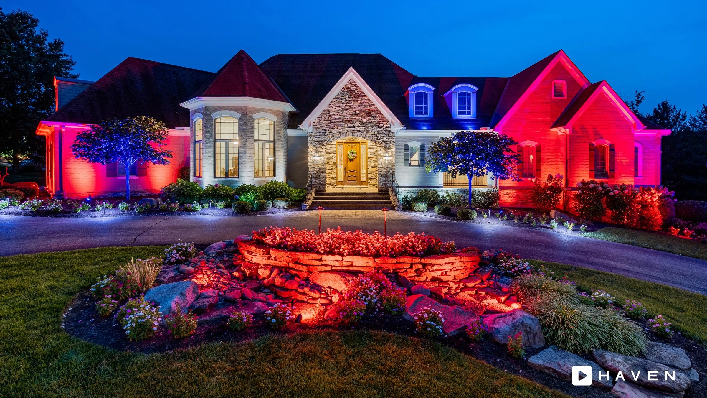 Nighttime view of a large house decorated with vibrant red, blue, and purple exterior lighting, surrounded by well-maintained garden beds, trees, and flowering plants.