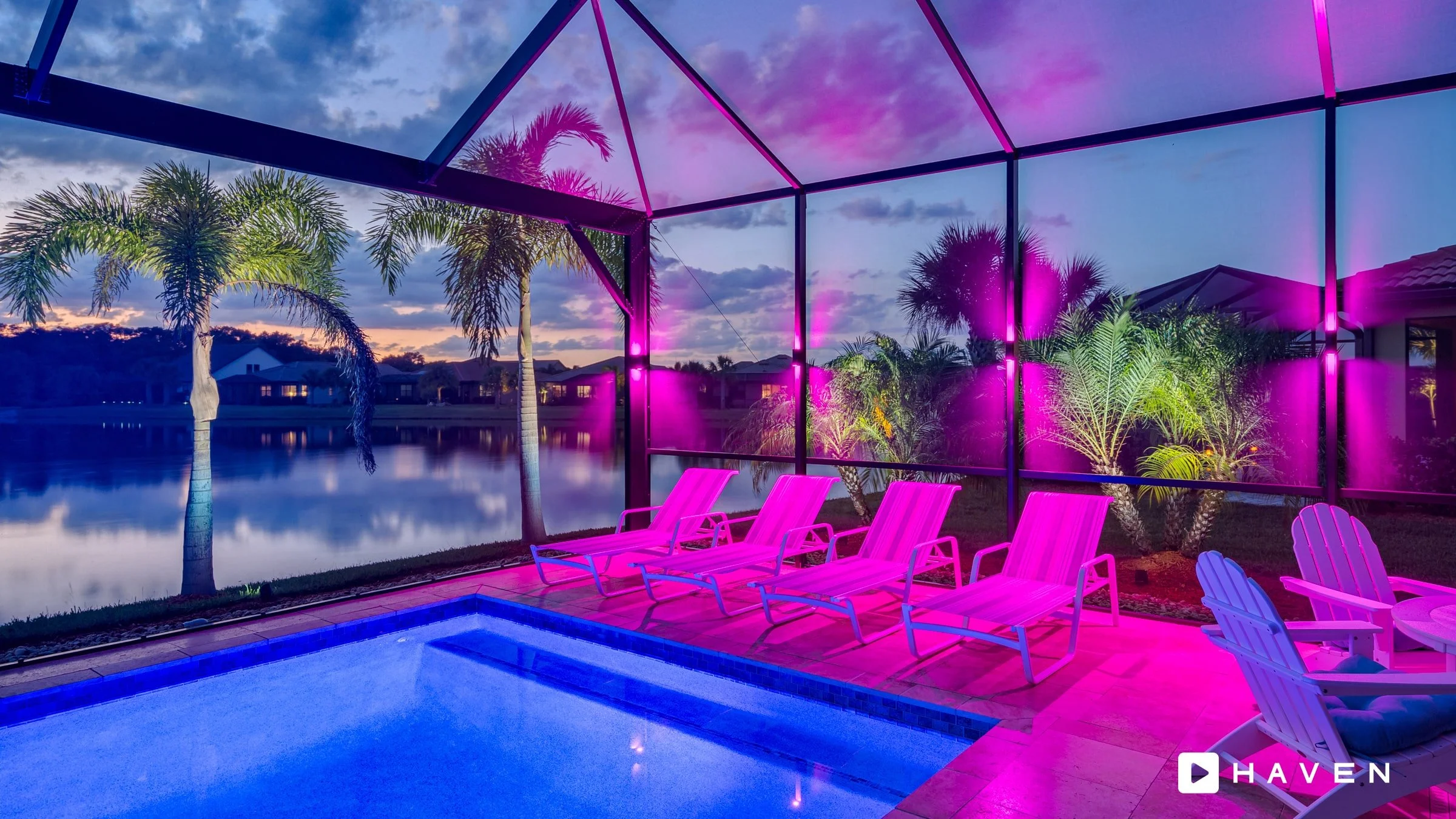 A backyard pool area at dusk with palm trees, smart landscape lighting, lounge chairs, and a view of a pond or lake with houses in the background.