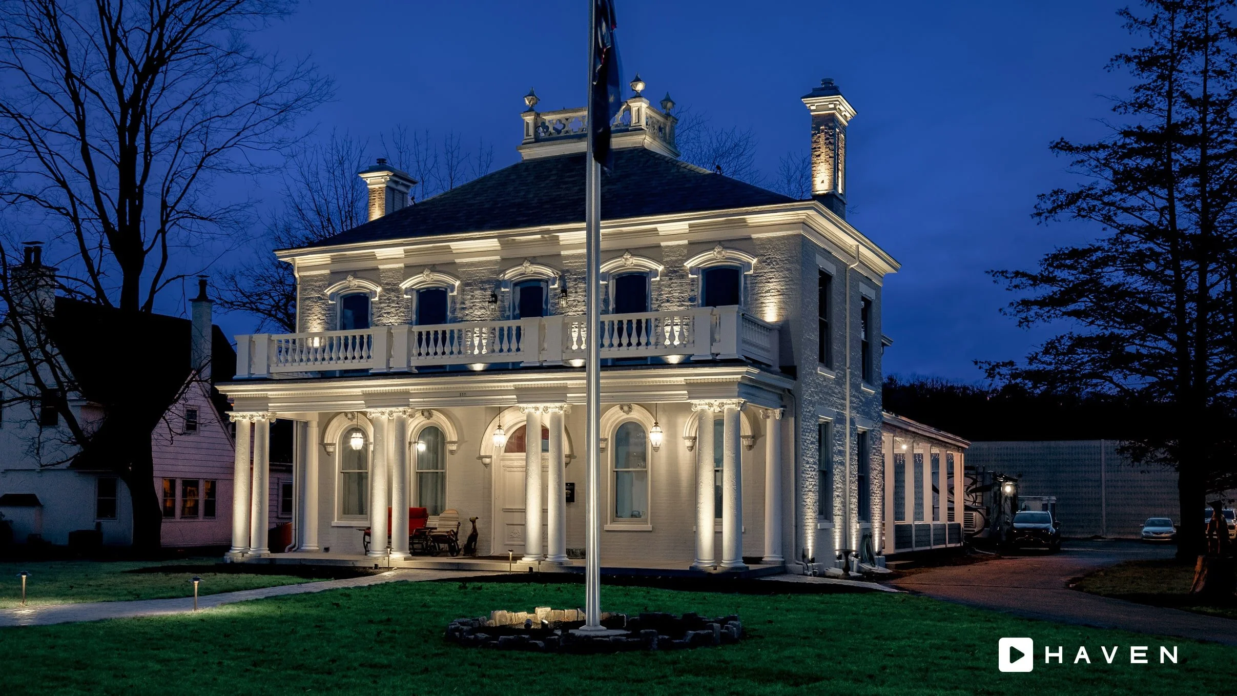 A historic white two-story house illuminated at night, with a flagpole in front and leafless trees in the background.