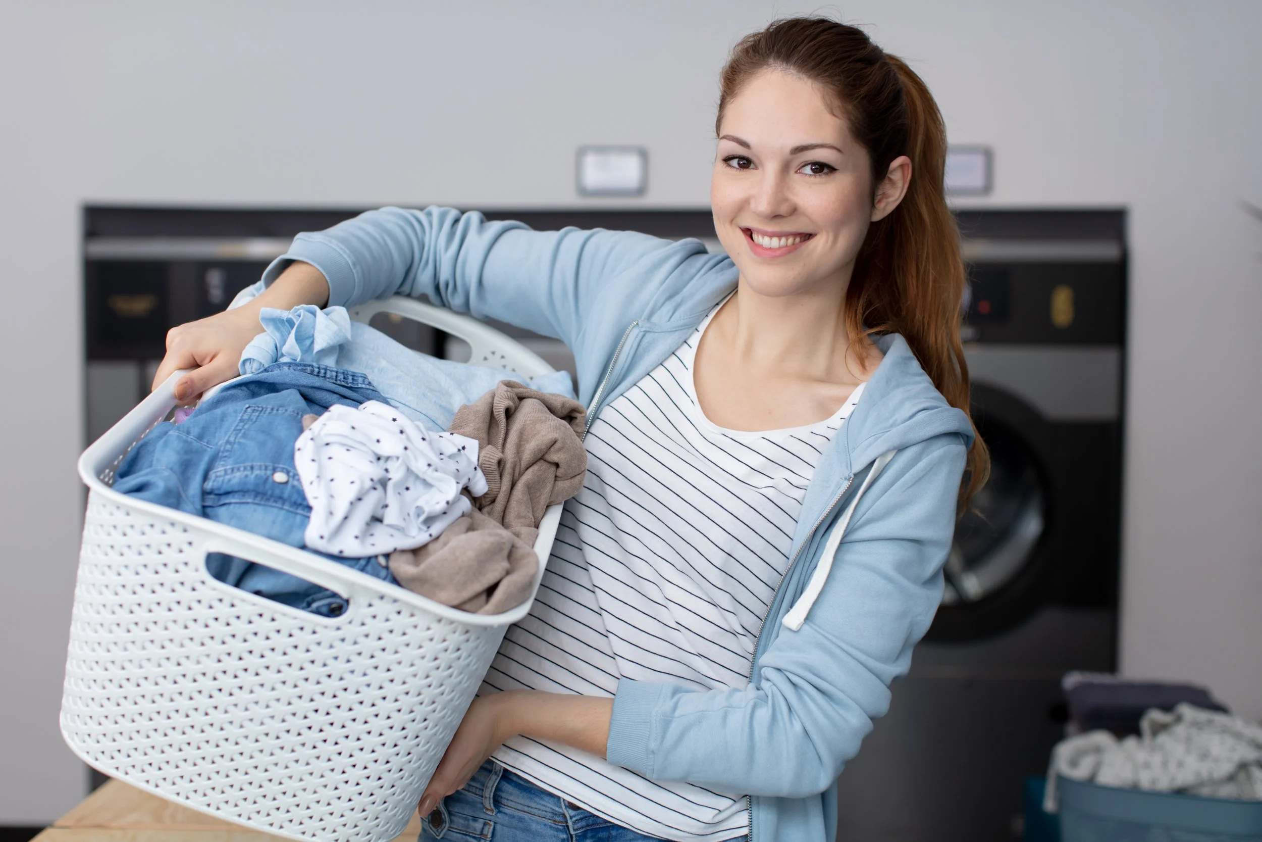 A young woman with light skin, brown hair tied back, and wearing a light blue hoodie and striped shirt, is smiling while holding a laundry basket overflowing with clothes in a laundry room.
