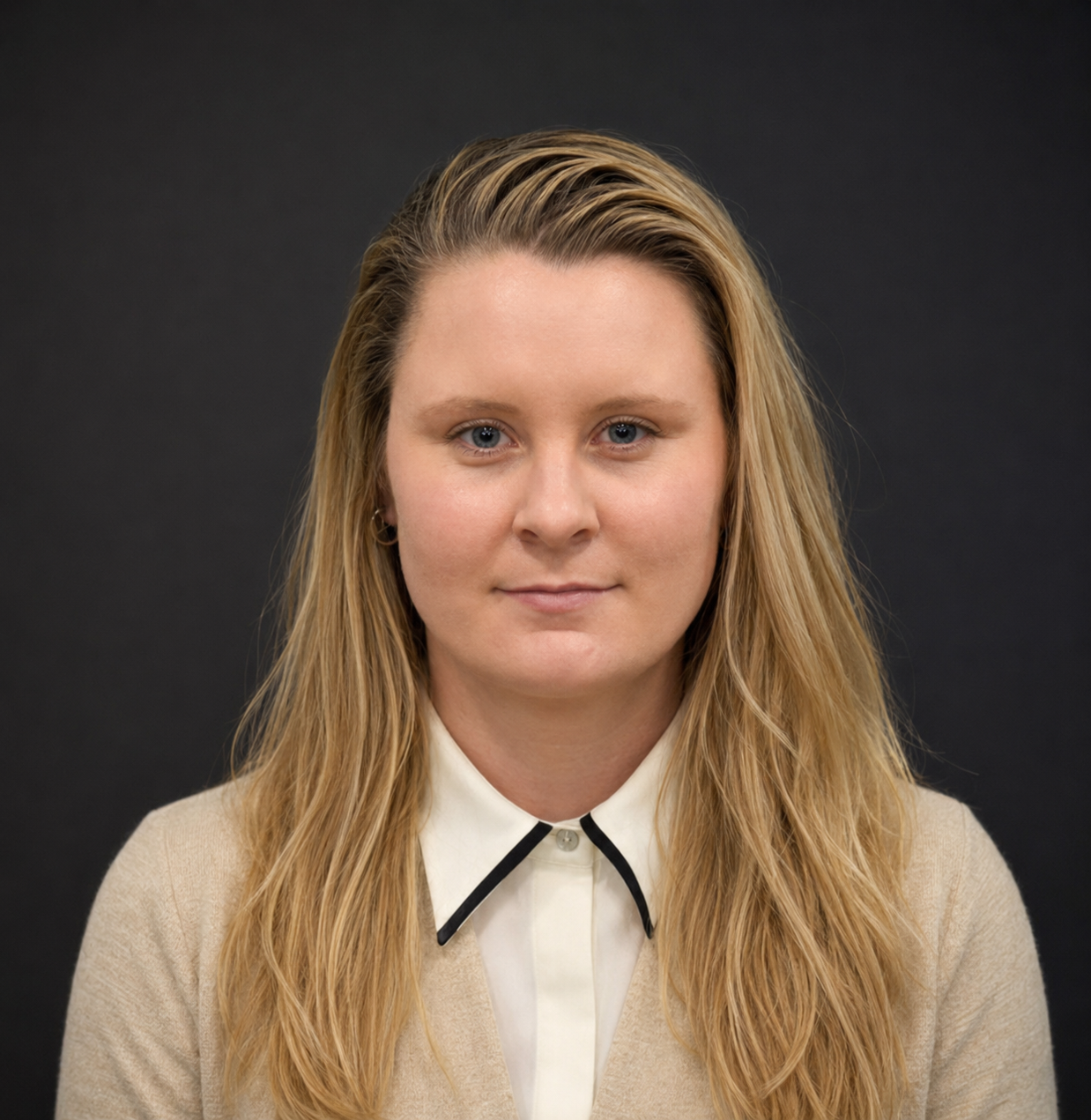 Portrait of a young woman with long blonde hair, blue eyes, and fair skin, wearing a beige sweater over a white collared shirt with black accent on the collar, against a dark plain background.