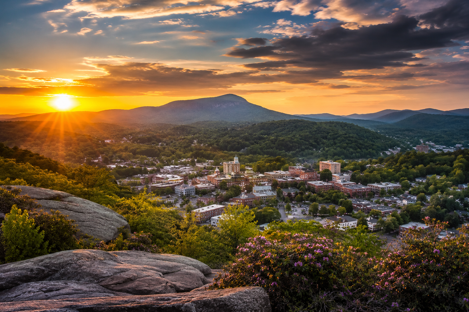 Sunset over a small town with mountains in the background, view from a rocky overlook with greenery and pink flowers in the foreground.