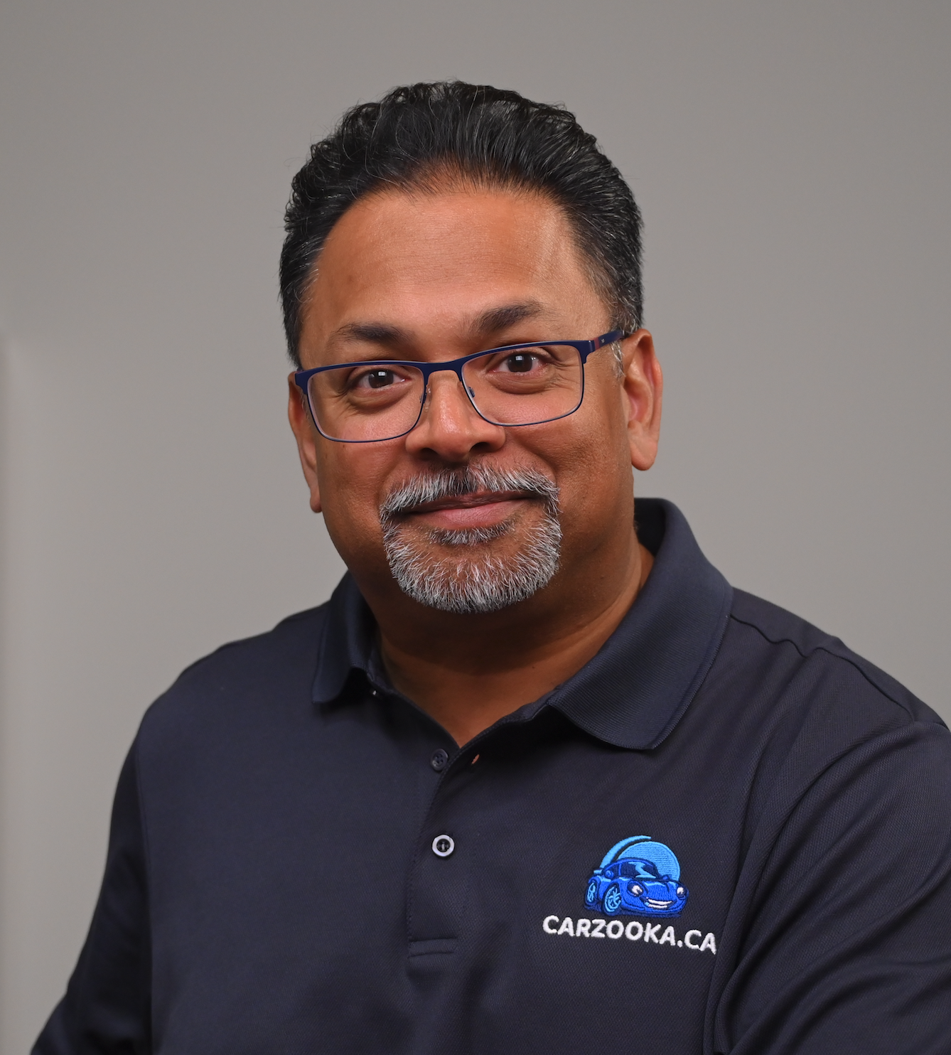 A man with glasses, graying hair, and a goatee, smiling, wearing a navy blue polo shirt with a Carzooka logo.