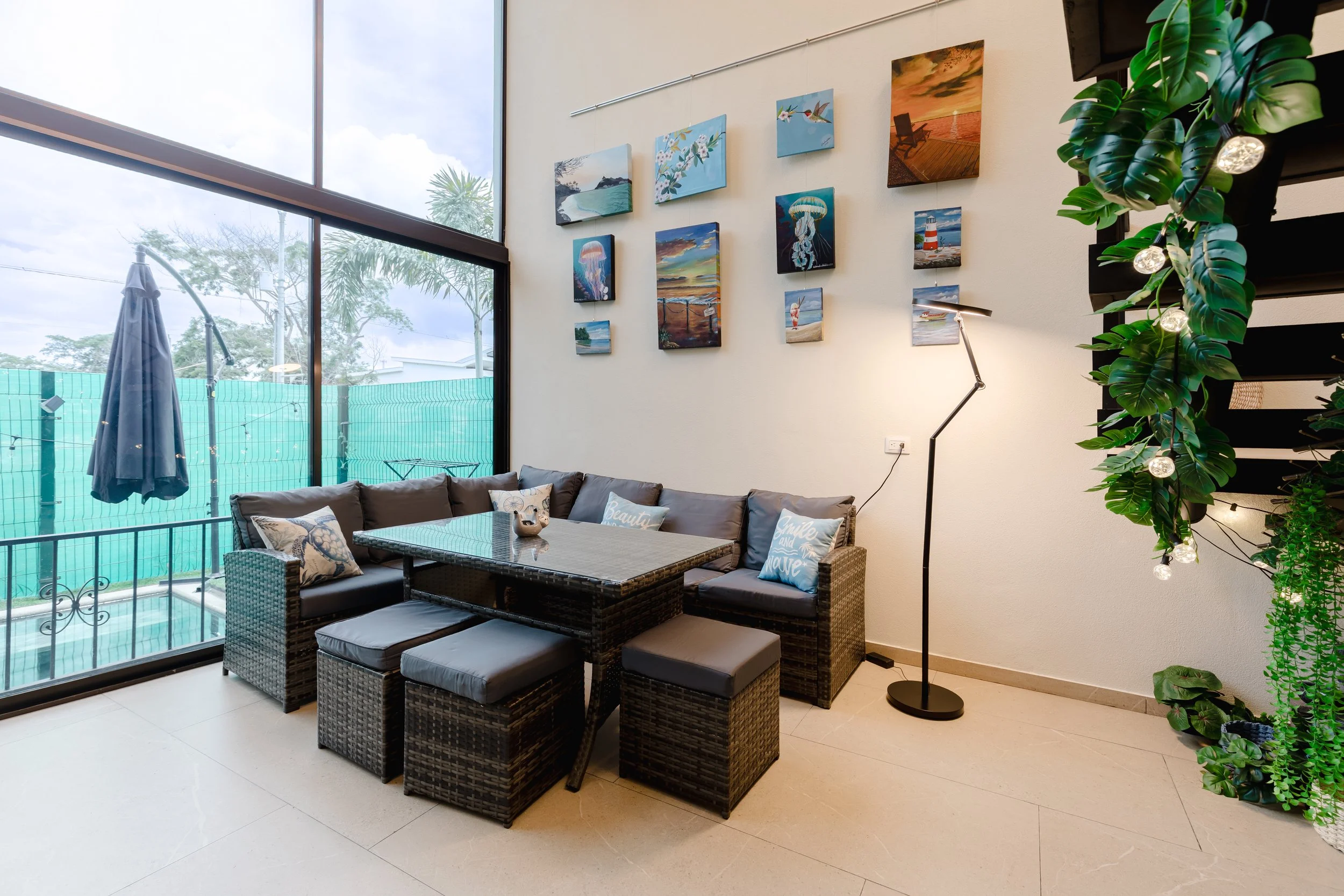 Indoor lounge area with large floor-to-ceiling windows showing outdoor trees and a patio umbrella, decorated with framed beach and nature artwork, a wicker dining set with cushioned seats and decorative pillows, a tall indoor plant with string lights, and a modern black floor lamp.