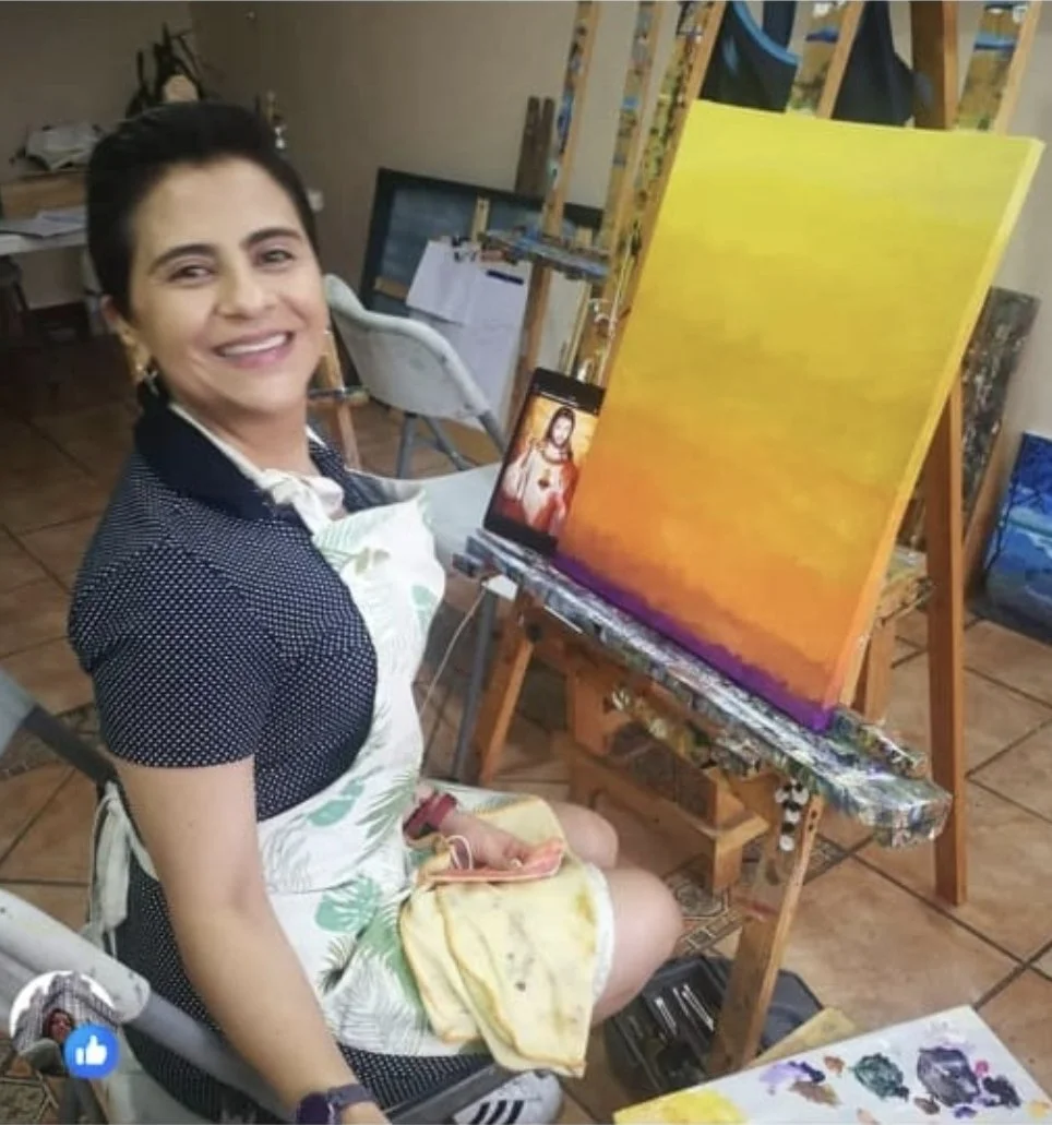 Woman sitting in an art studio, smiling, with an unfinished gradient sunset painting on an easel in front of her. The background shows art supplies and other paintings.