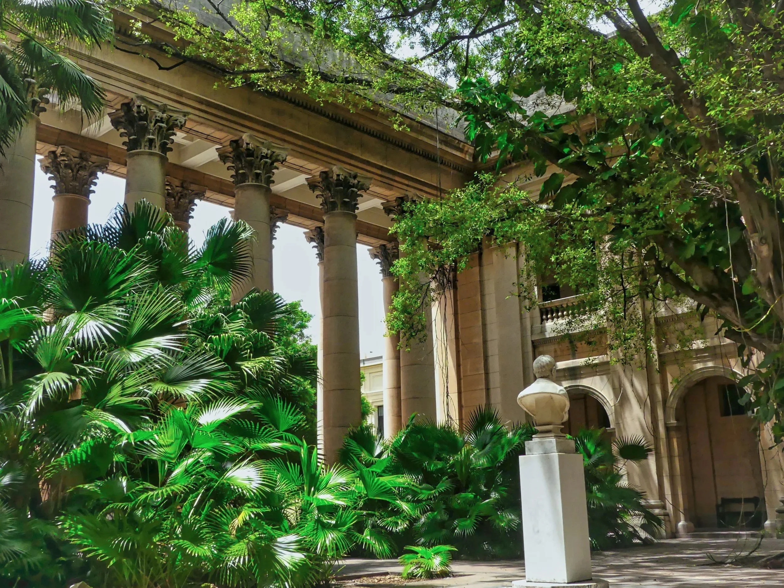 A view of an outdoor architectural structure with large stone columns and a decorative entablature, surrounded by lush green trees and plants.