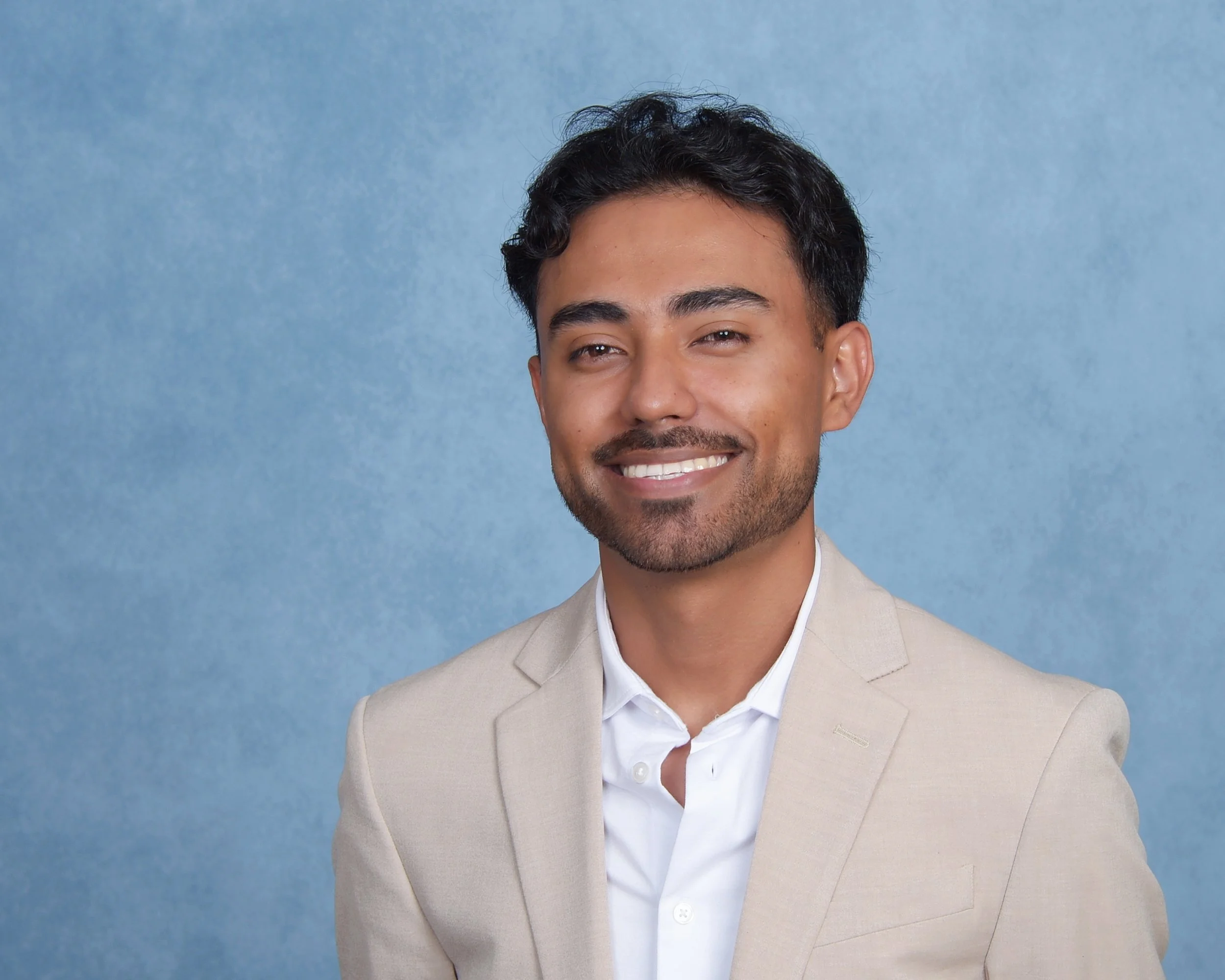 A young man with dark curly hair and a beard, wearing a light-colored suit jacket and a white shirt, smiling in front of a blue background.