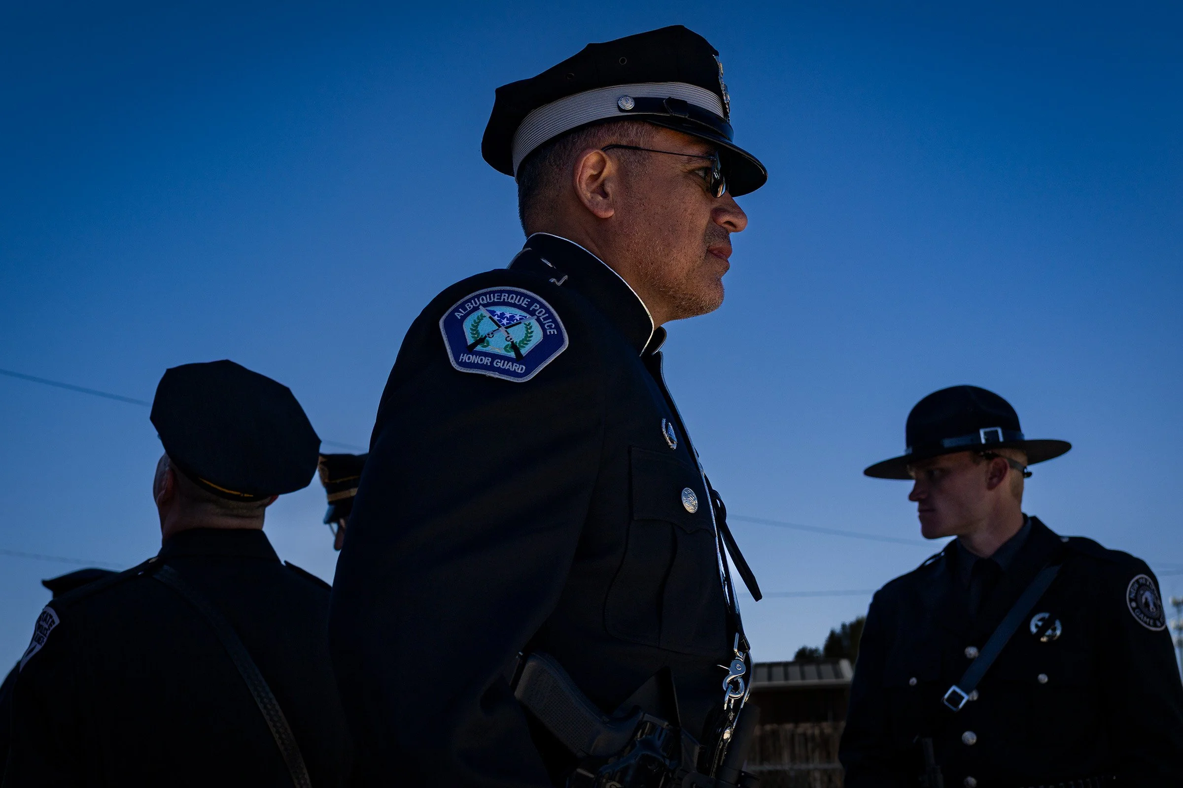 A group of police officers wearing dark uniforms and hats, standing outdoors during dusk or dawn, with a clear sky and some trees in the background.