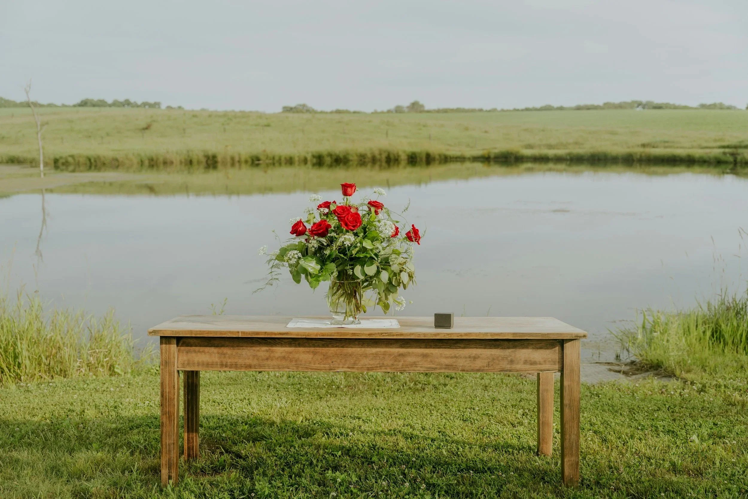 A wooden table with a bouquet of red roses and white flowers in a glass vase, placed outdoors by a lake with a green grassy area and a distant hill in the background.