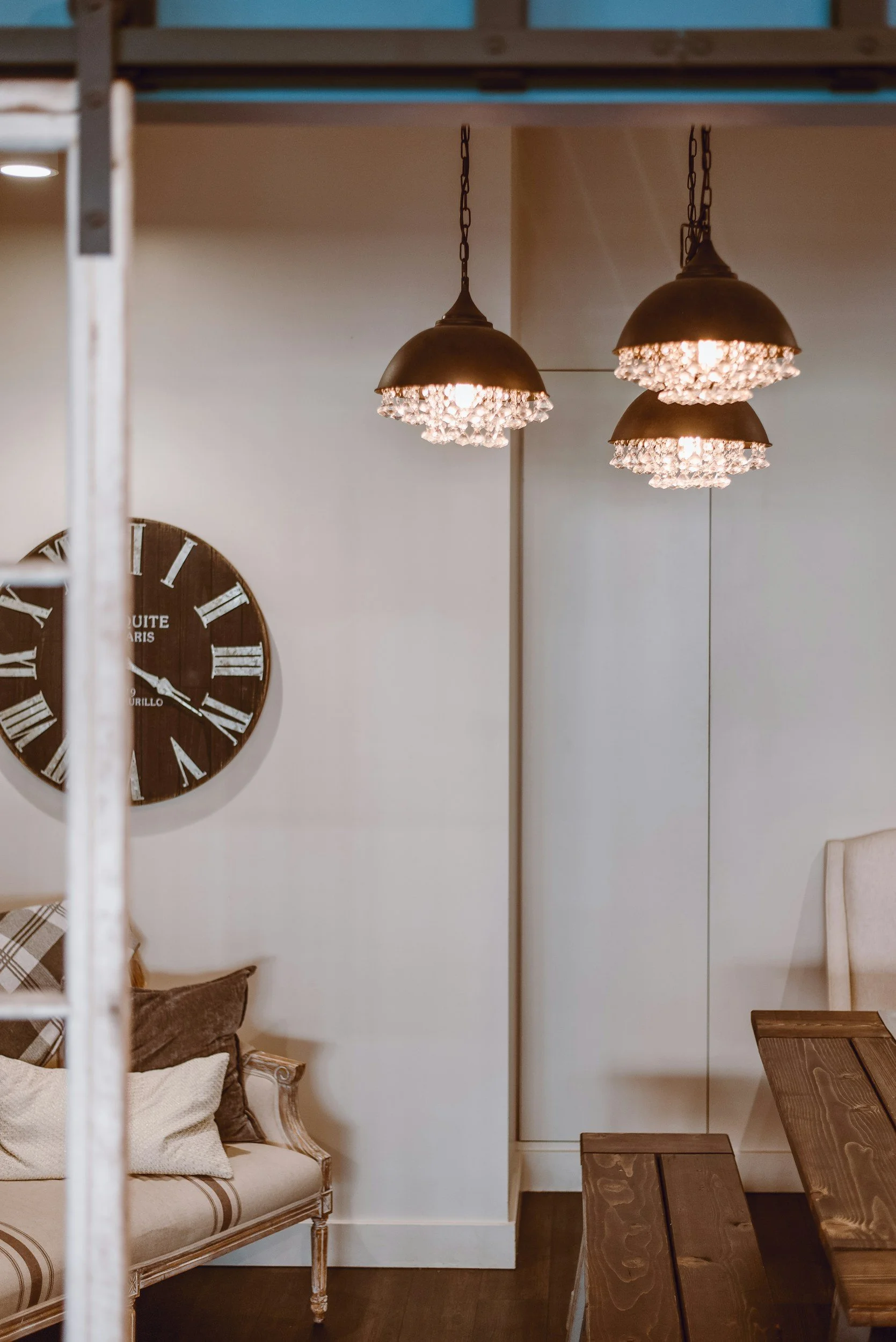 Interior view of a room with a beige sofa, cushions, a hanging wall clock, wooden dining table, pendant lights, and a partition wall.