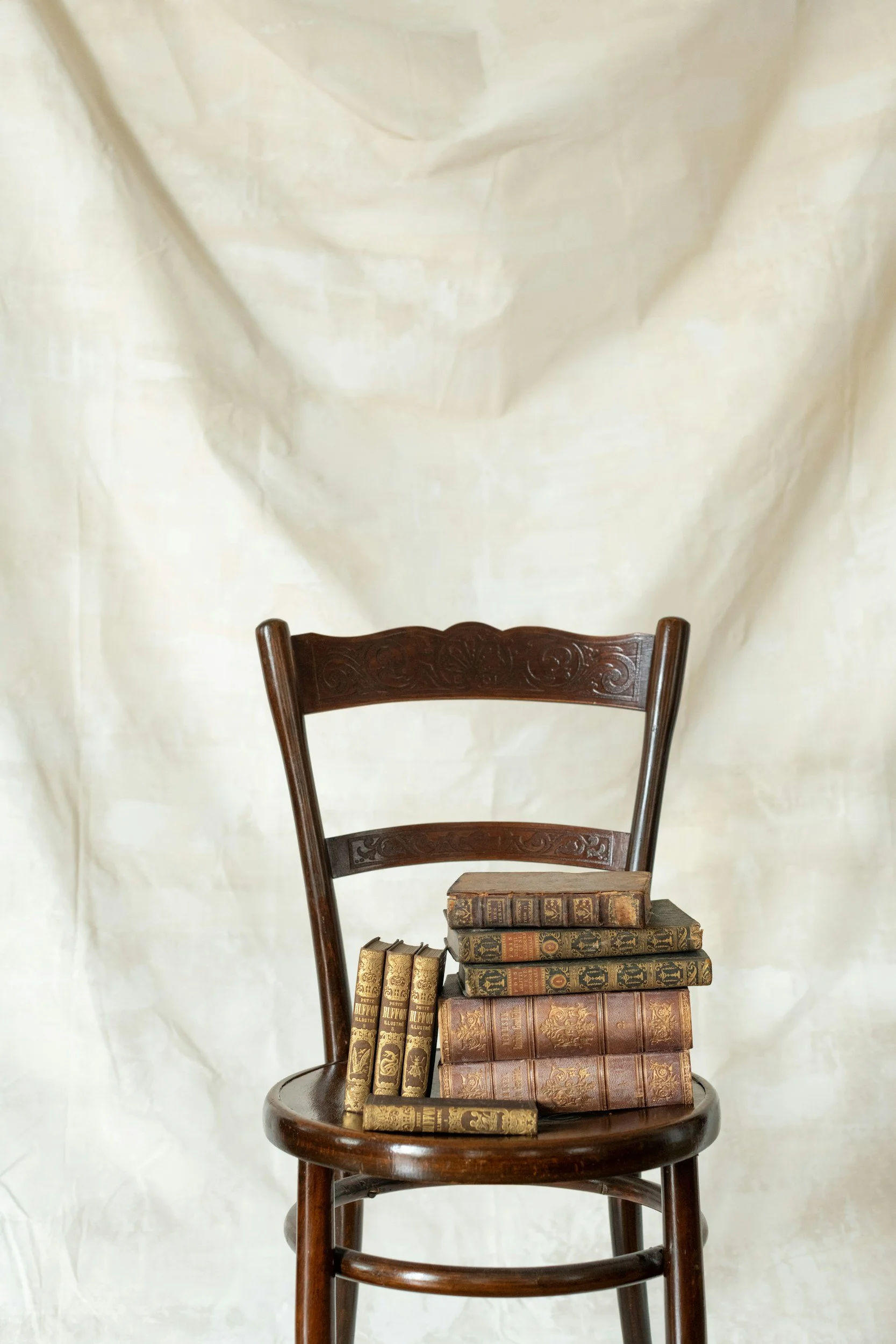 A wooden chair with a stack of old antique books placed on the seat, against a plain light-colored fabric background.