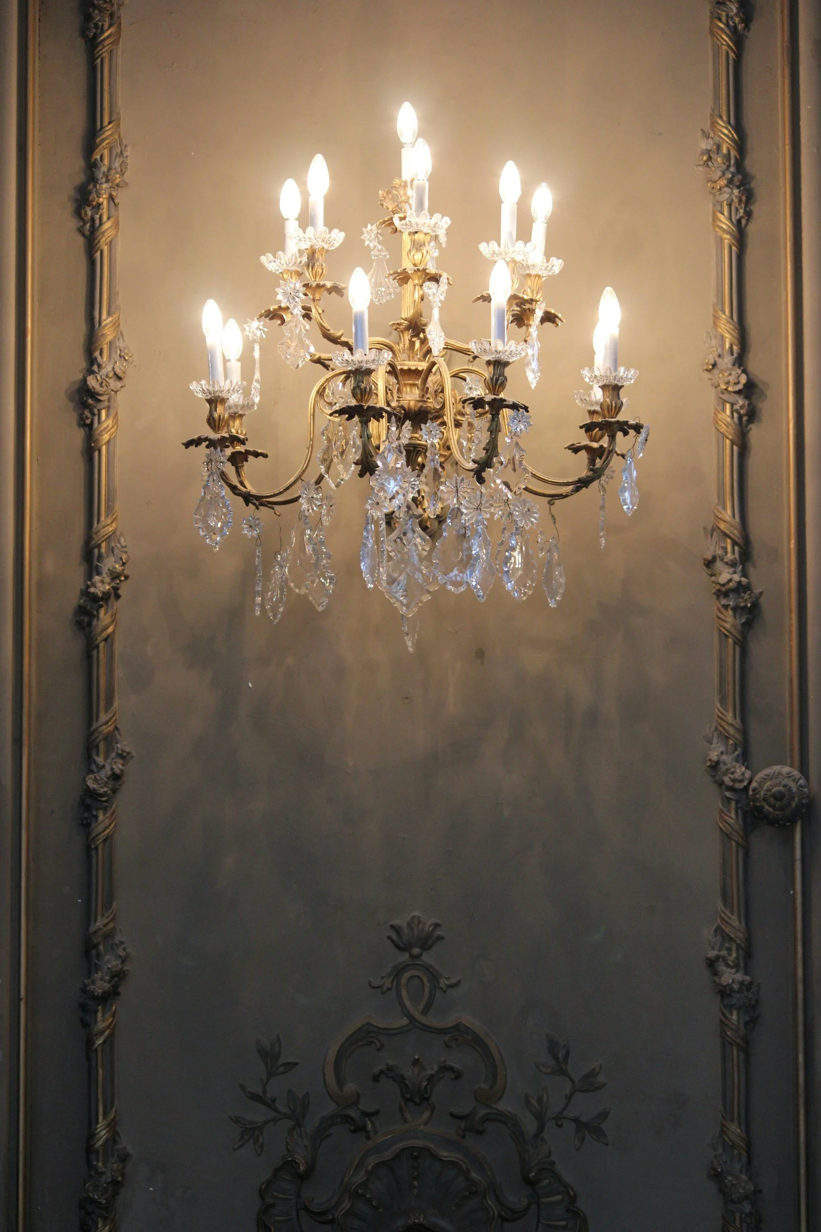 A luxurious chandelier with multiple candle-shaped lights, adorned with crystal decorations, hanging against a classical-style wall with ornate molding.