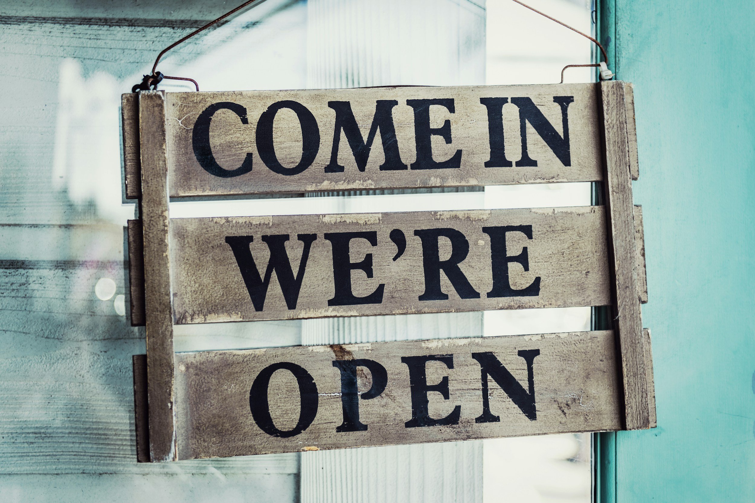Wooden sign with the words 'Come in We're Open' hanging on a hook outside a building.