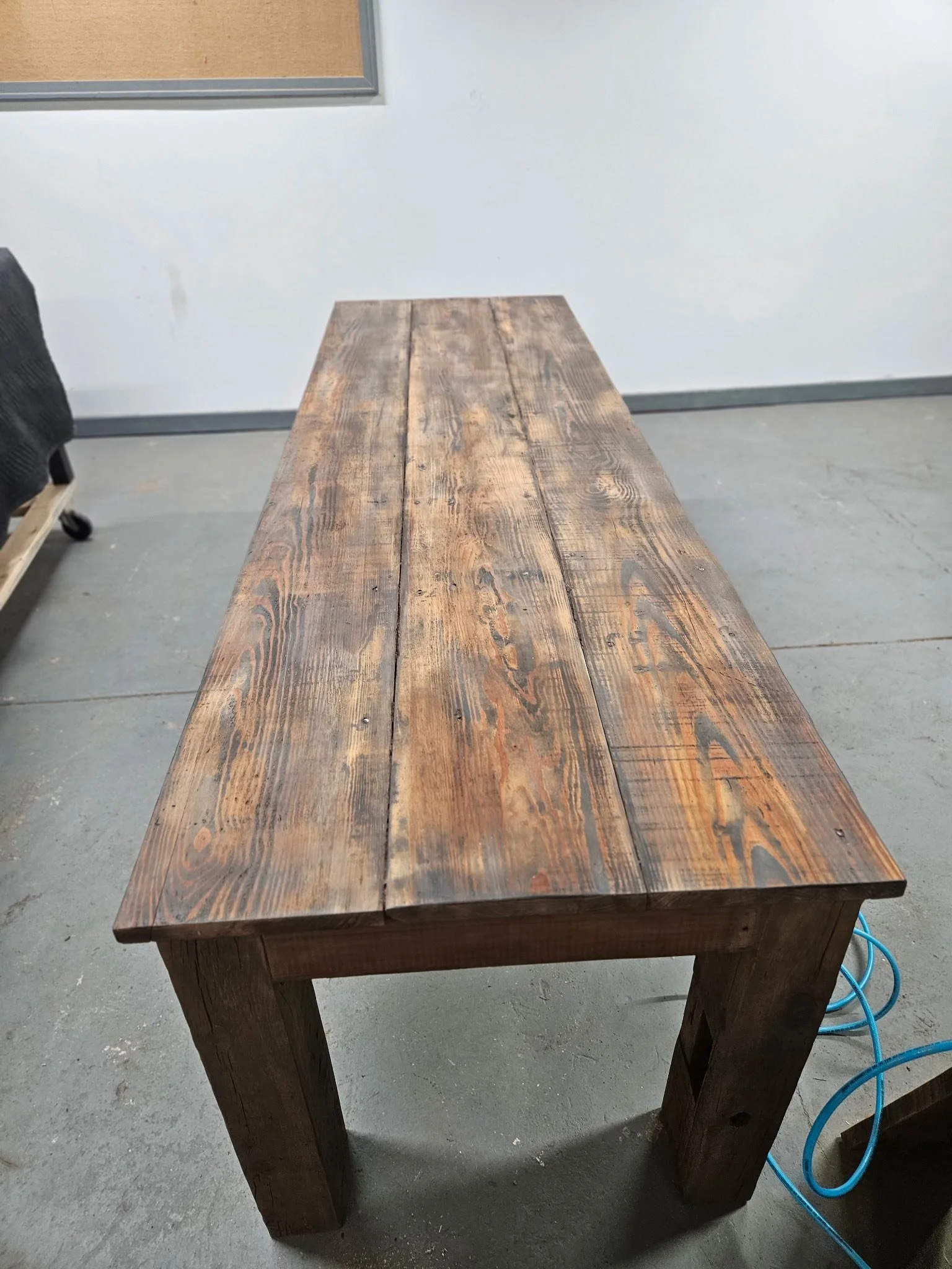 A wooden table with a rustic, stained surface positioned in a room with a plain white wall, a cork board on the wall, and a blue power cord on the floor.
