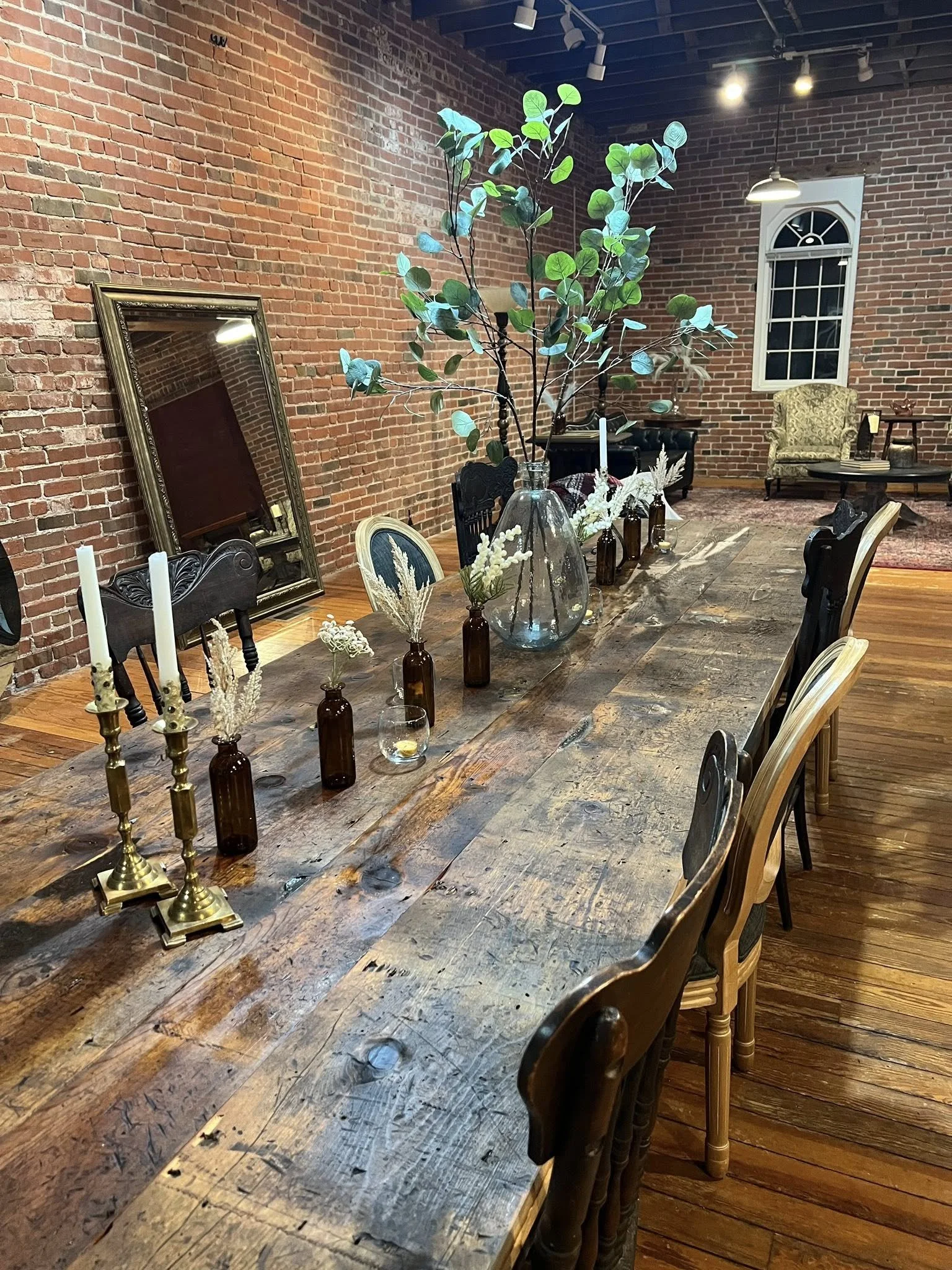 A rustic dining room with a long wooden table decorated with candles and vases, a large mirror leaning against a brick wall, and a cozy seating area with an armchair and a window.