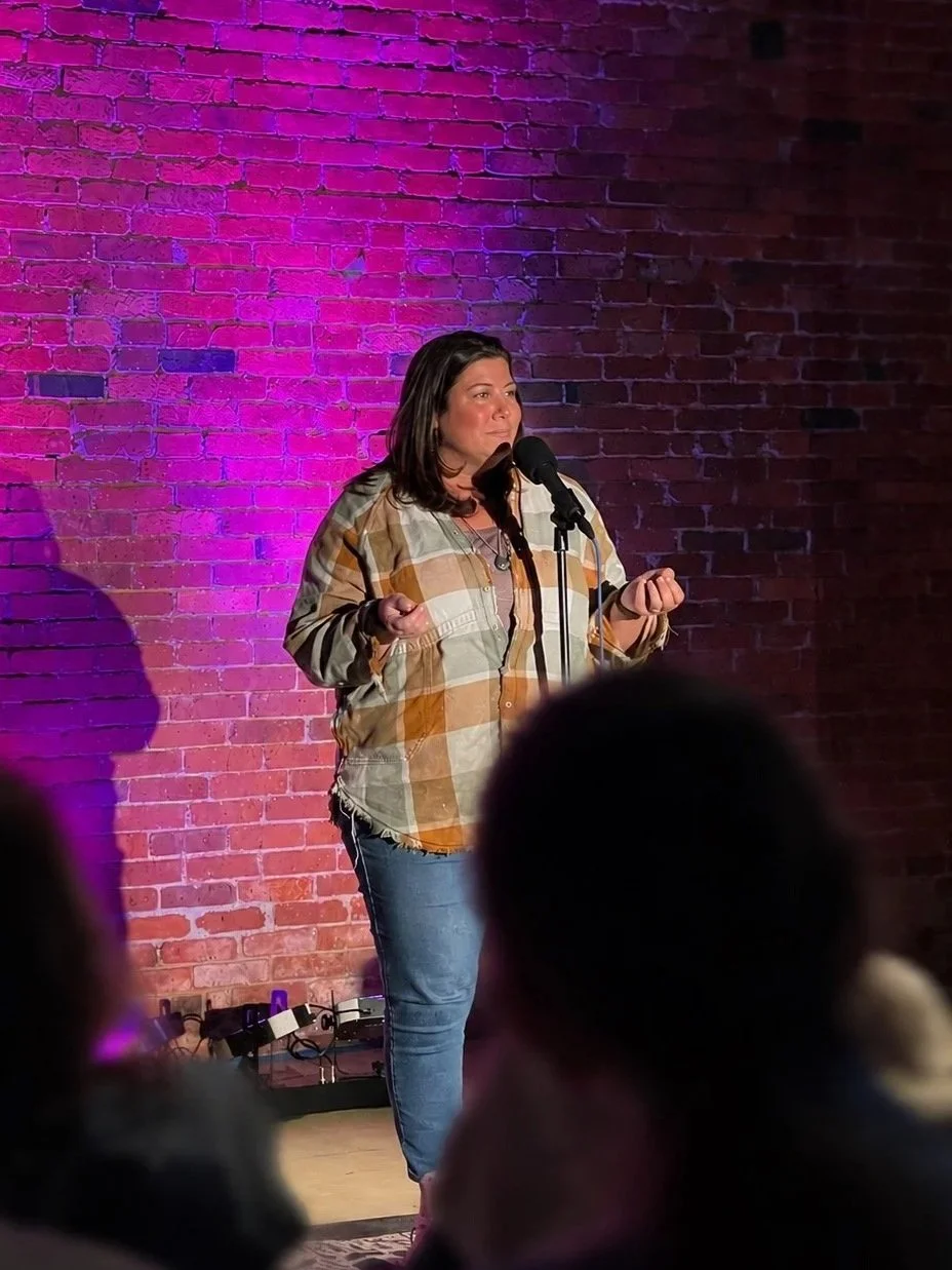 A woman speaking into a microphone on stage against a brick wall with purple lighting, audience members visible in the foreground.