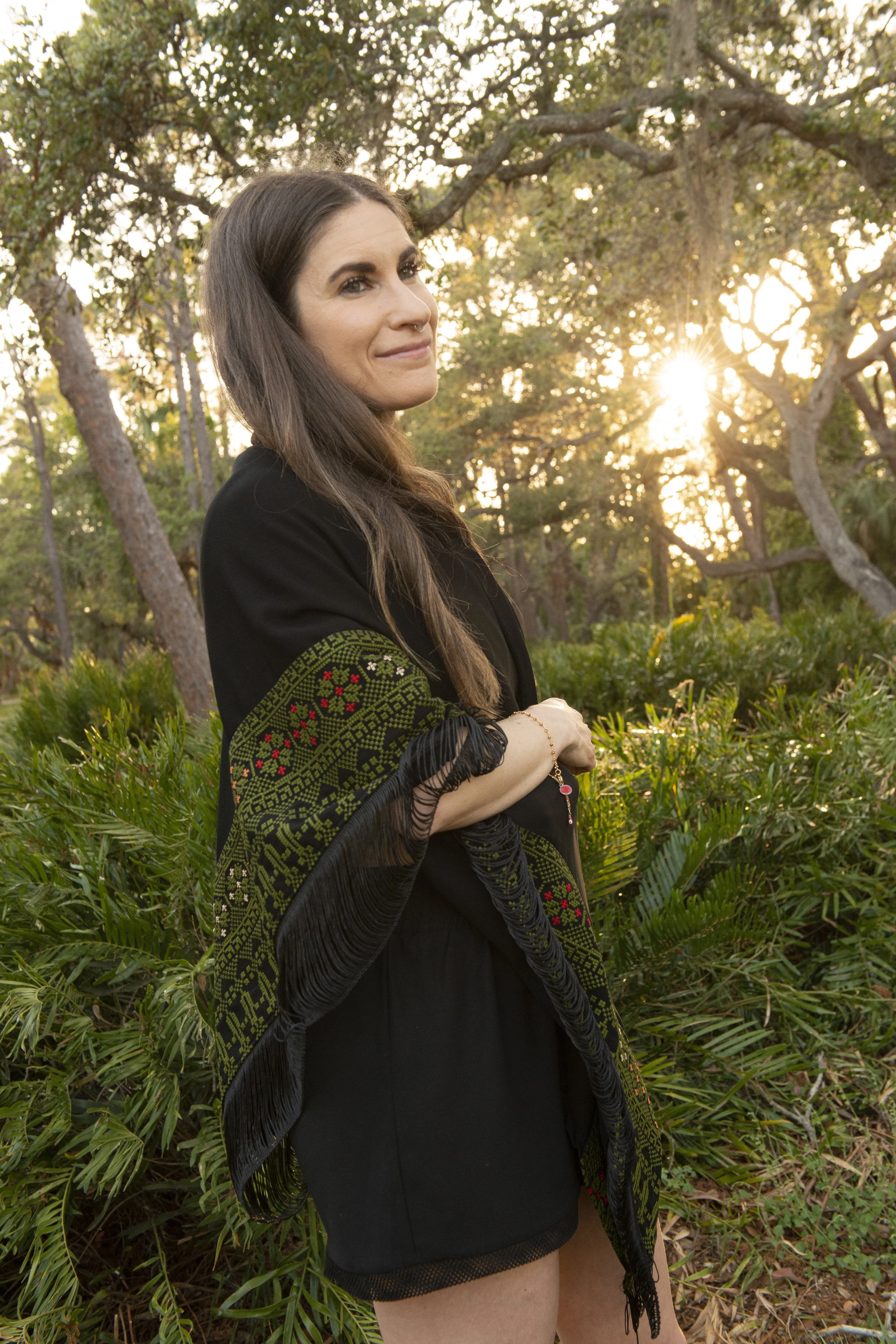 Amanda Elemental stands outdoors surrounded by greenery and trees, with sunlight filtering through the branches at sunset.