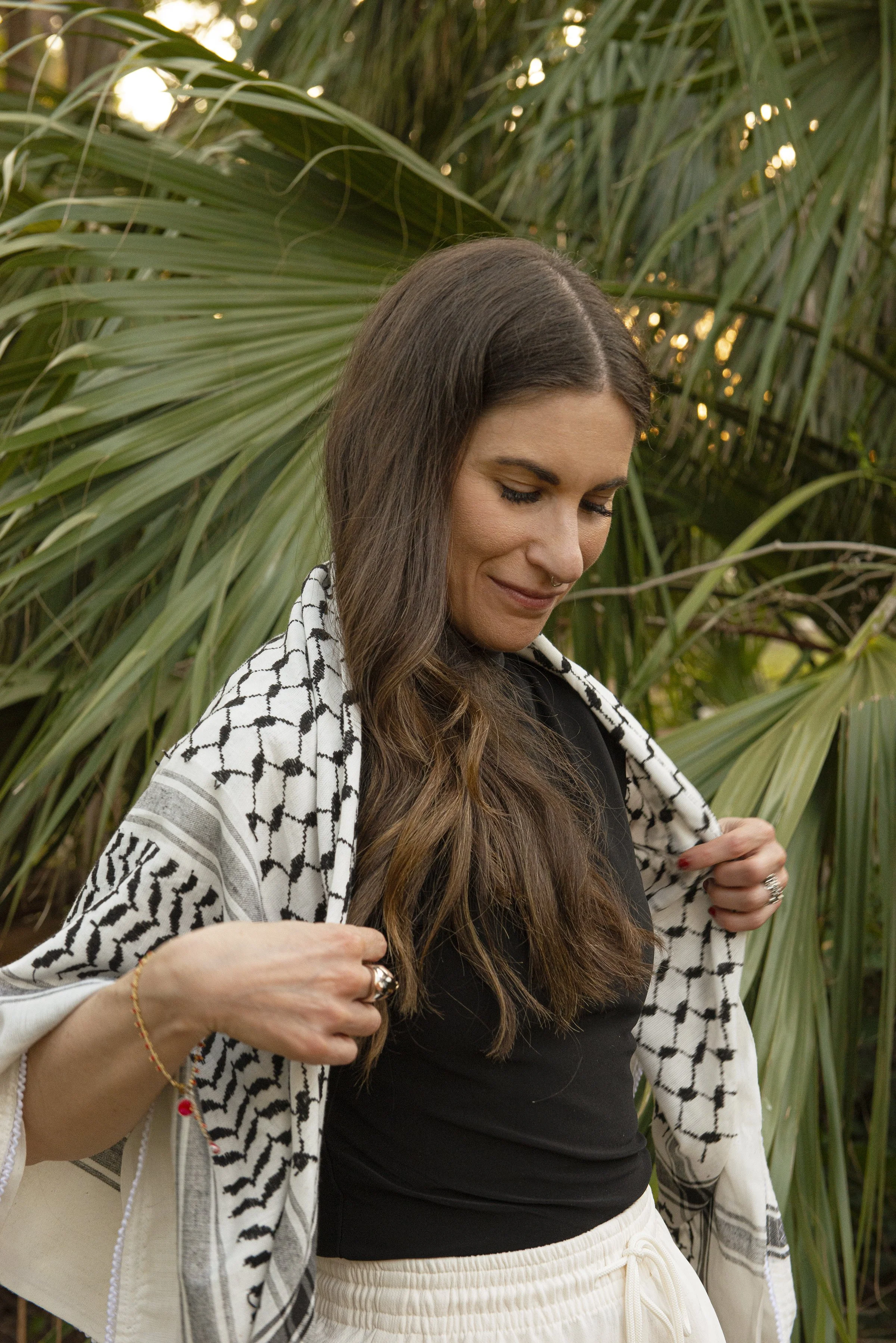 A woman with long brown hair, wearing a black top and white sweatpants, stands outdoors with large green palm leaves in the background. She has a patterned black and white shawl over her shoulders and is smiling softly while looking downward, with her hands adjusting her clothing.