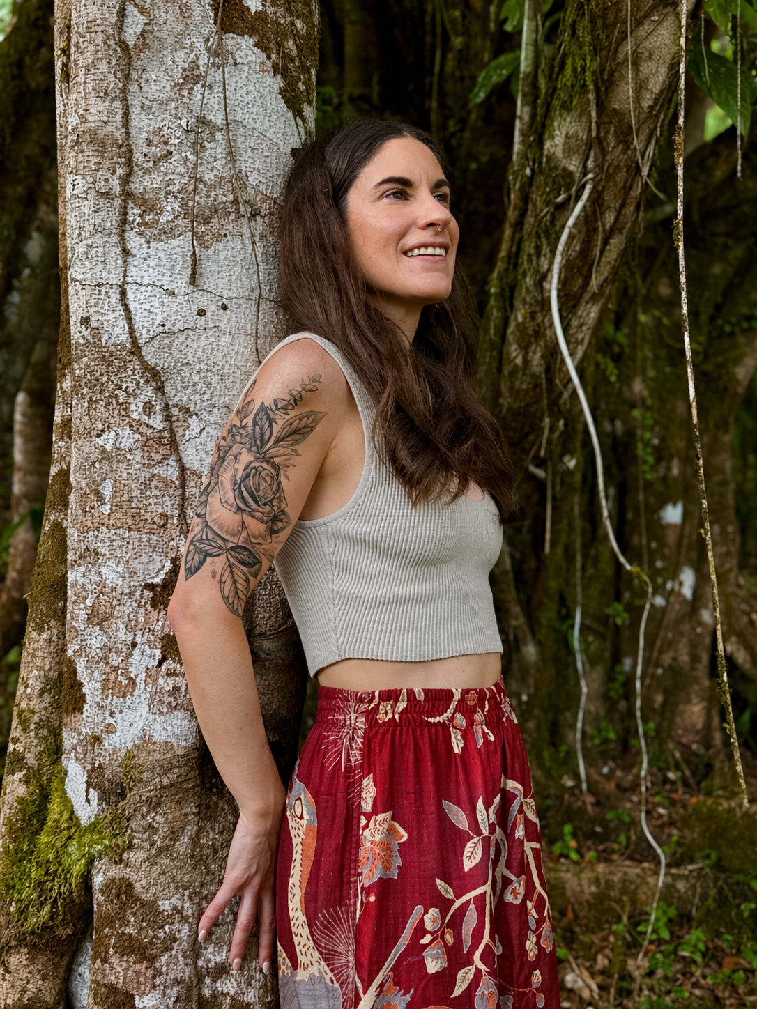 A woman with long dark hair and a tattoo of flowers on her arm, wearing a gray crop top and red patterned skirt, leaning against a tree in a forest.