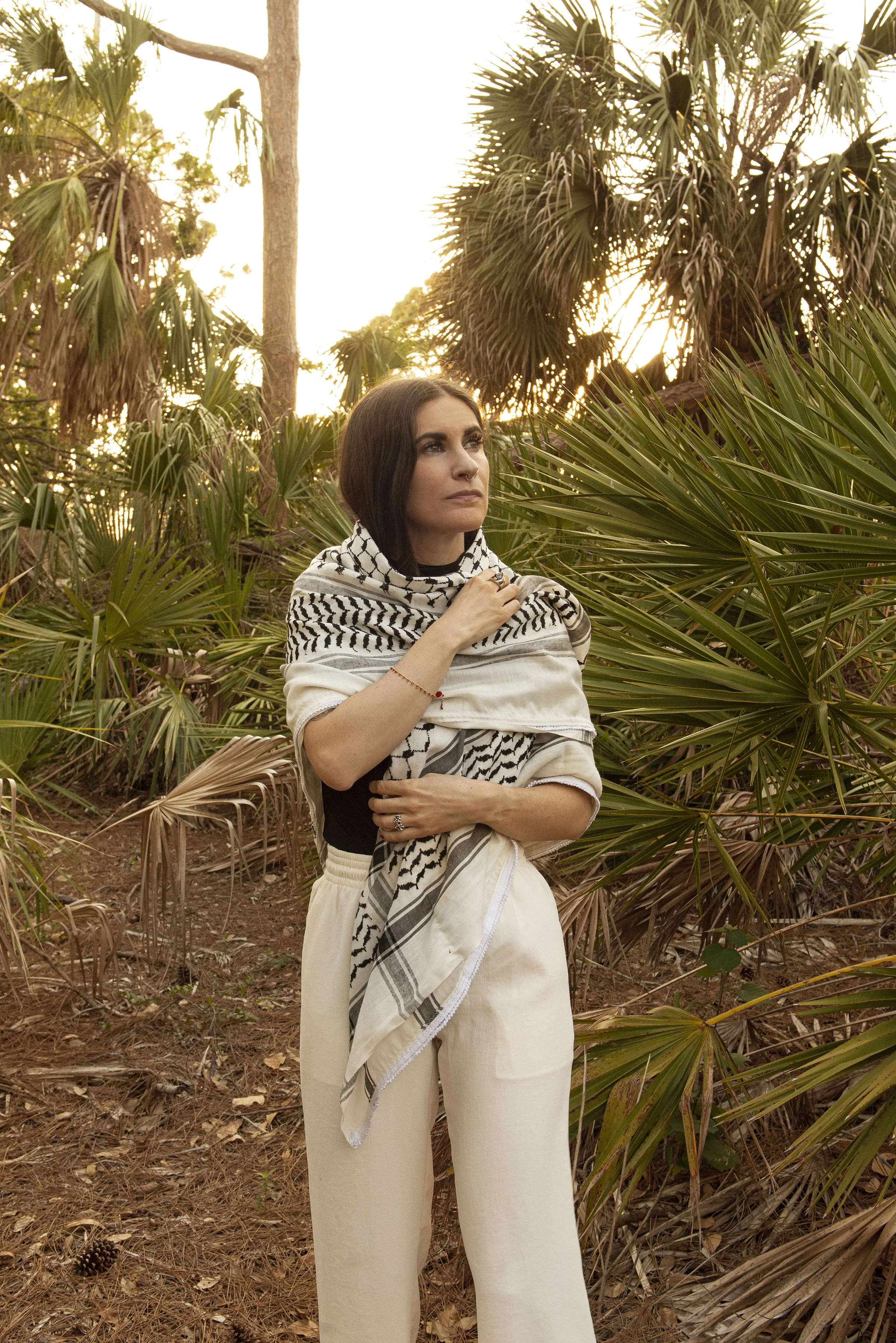 A woman with dark hair, wearing a black and white patterned scarf, stands amidst lush green palm trees and tropical plants at sunset.