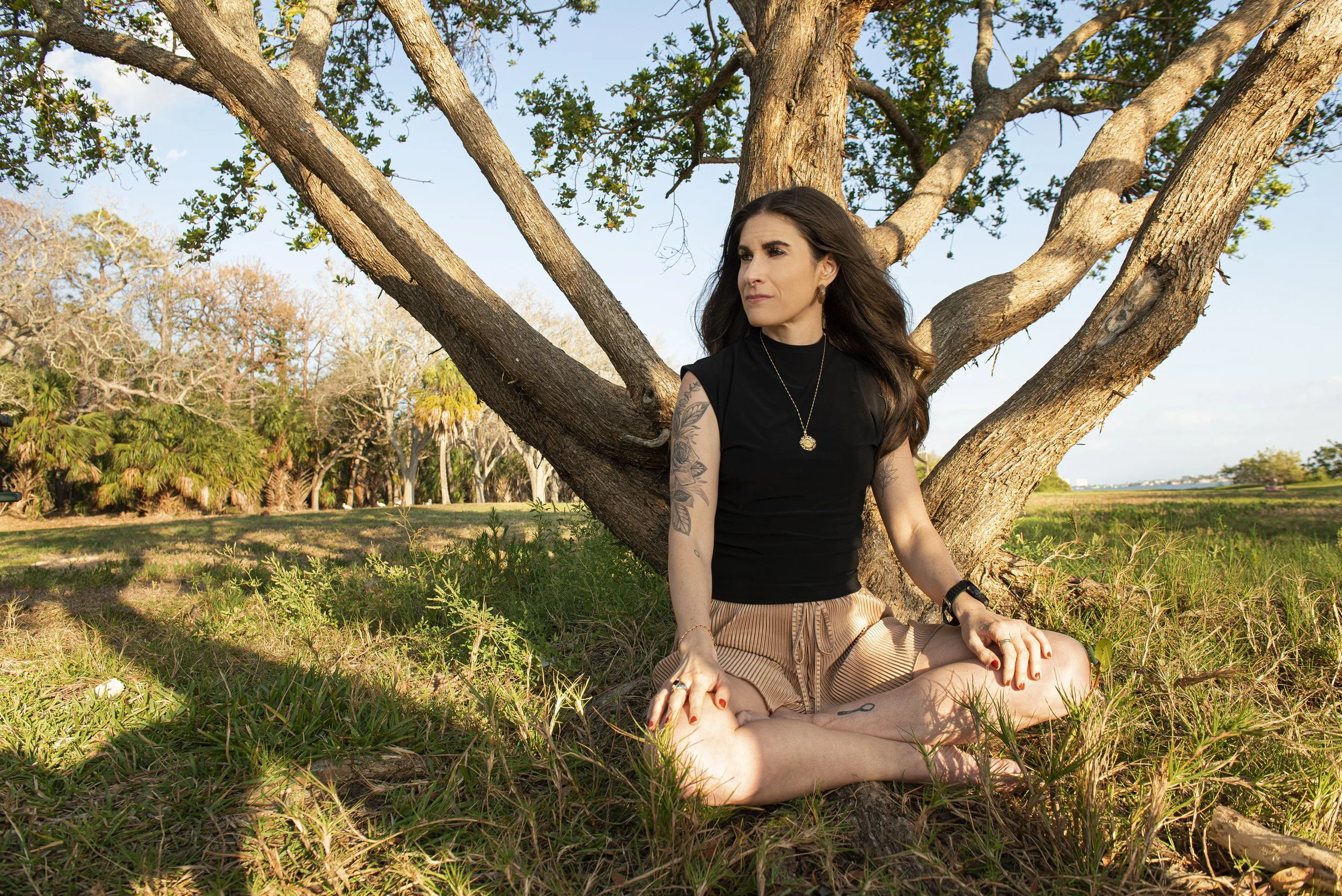 A woman with long dark hair and tattoos sits cross-legged under a tree in a park on a sunny day, wearing a black top and beige shorts, with lush green grass and trees around her.
