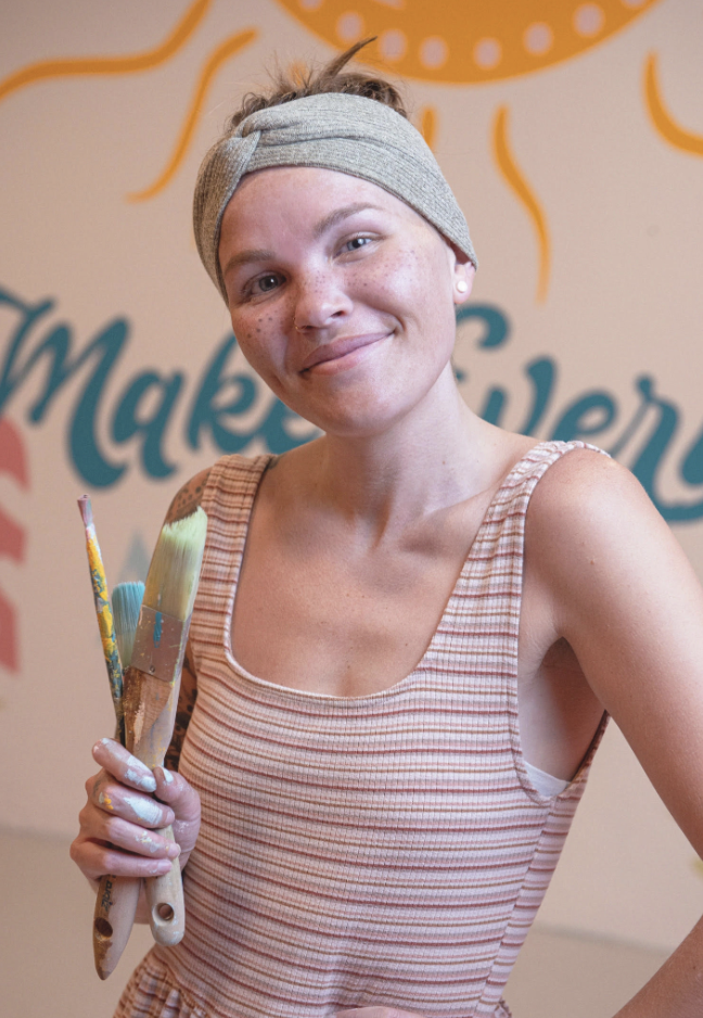 Young woman with short hair and freckles, wearing a gray headband and sleeveless striped shirt, smiling, holding paintbrushes, standing in front of a colorful mural with the partially visible word 'Maker'.
