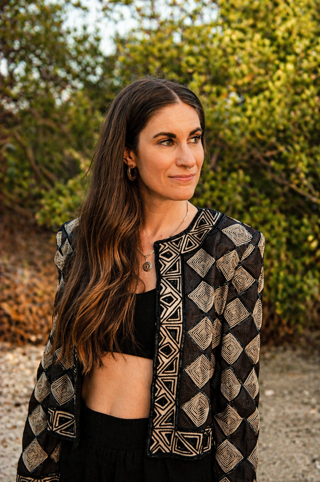 Amanda Elemental stands outdoors in front of green foliage. She wears a black crop top, black pants, a patterned jacket with geometric designs, earrings, and layered necklaces.