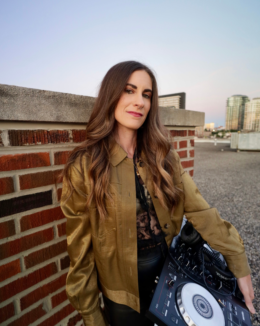Amanda Elemental leaning against a brick wall on a rooftop, holding a DJ controller, with city buildings in the background during sunset.