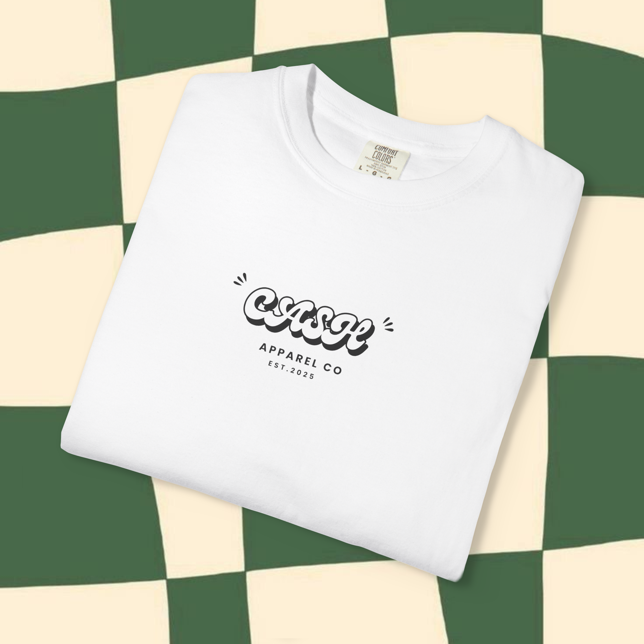 Close-up of a white T-shirt with the Cash Apparel C. logo, lying on a green and cream checkerboard patterned floor.