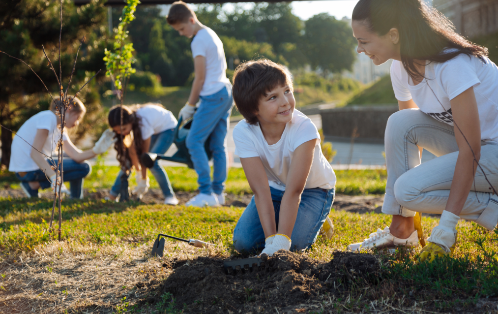 Children and a woman planting a tree outdoors on a sunny day