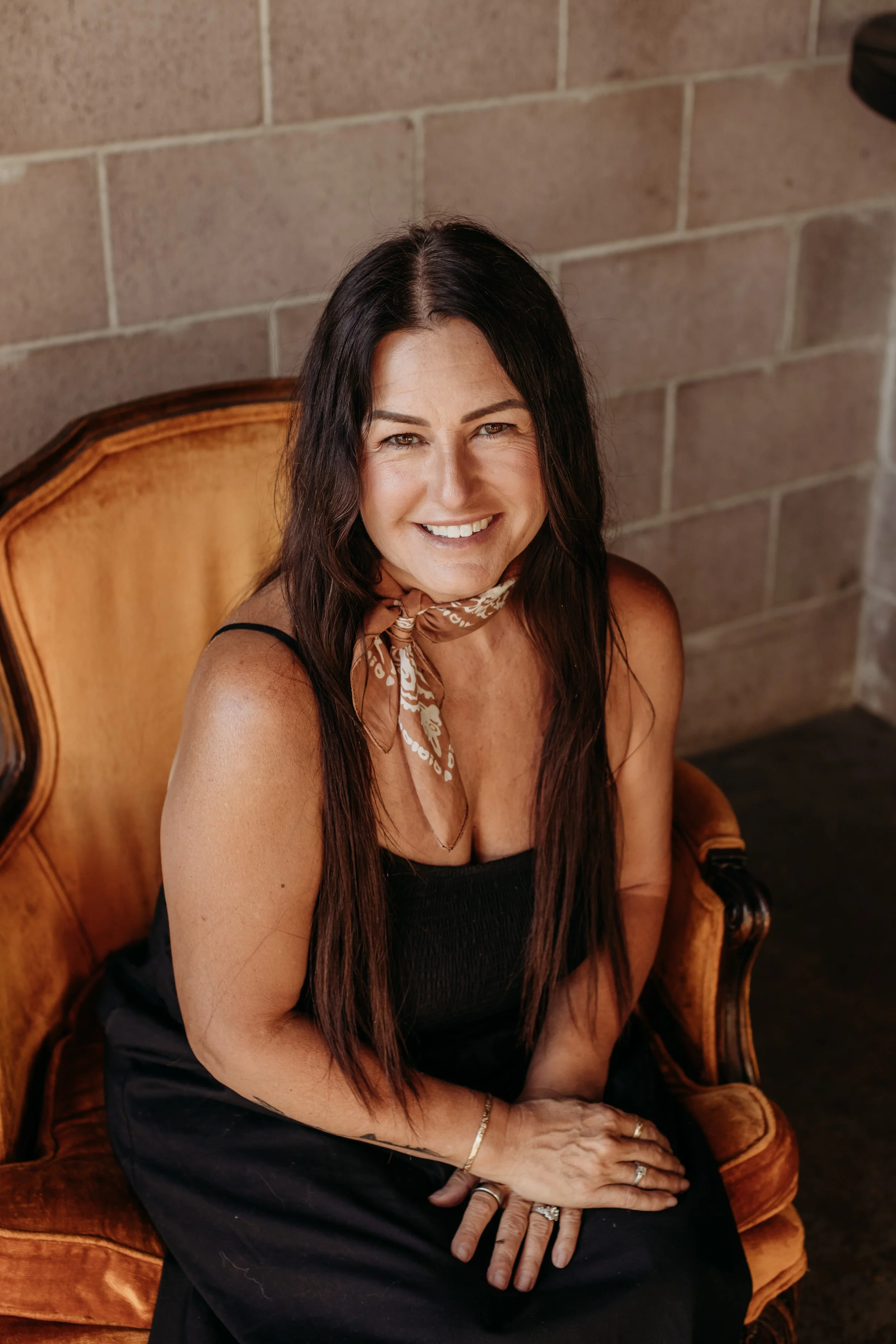 A woman smiling, sitting on a vintage wooden armchair with a high back, against a brick wall. She has long dark hair, wearing a black dress, a beige scarf with a white pattern, and jewelry including rings and a bracelet.