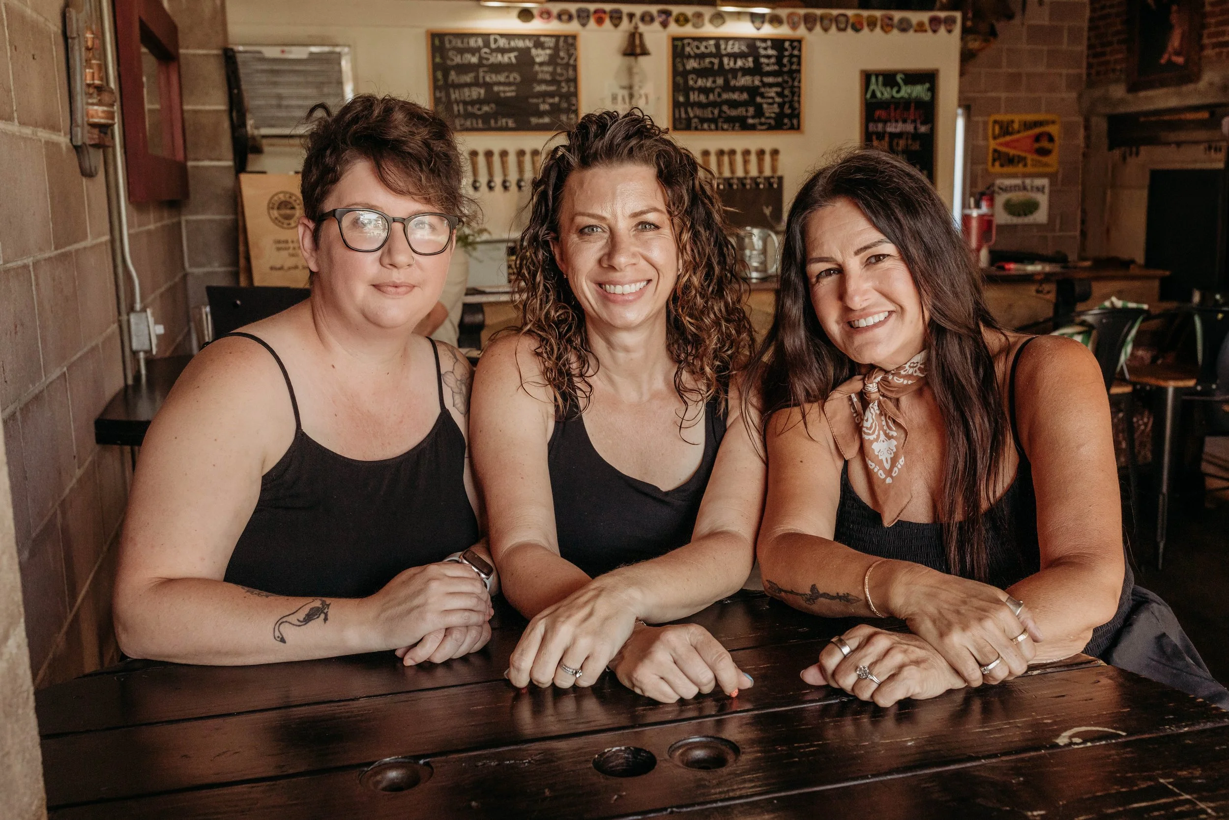 Three women sitting at a dark wooden table inside a restaurant or pub, smiling at the camera. The woman on the left has short, dark hair, glasses, and a tattoo on her left arm. The woman in the middle has curly brown hair and is wearing a black tank top. The woman on the right has long dark hair, a bandana around her neck, and is wearing a black top. In the background, there are chalkboard menus with food and drink options and various decorations on the wall.