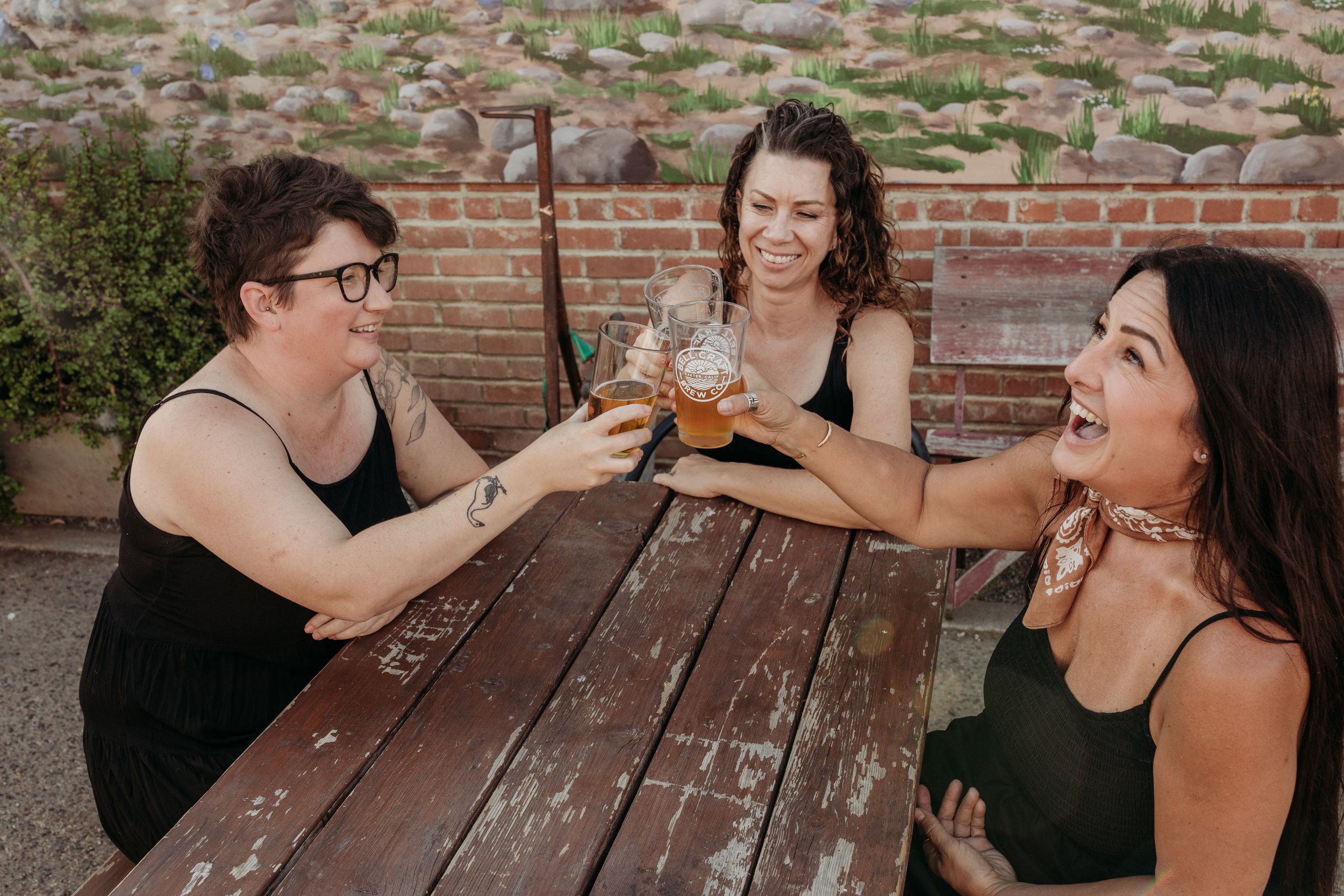 Three women sitting at a wooden picnic table outdoors, sharing drinks and smiling, with a brick wall and painted mural in the background.