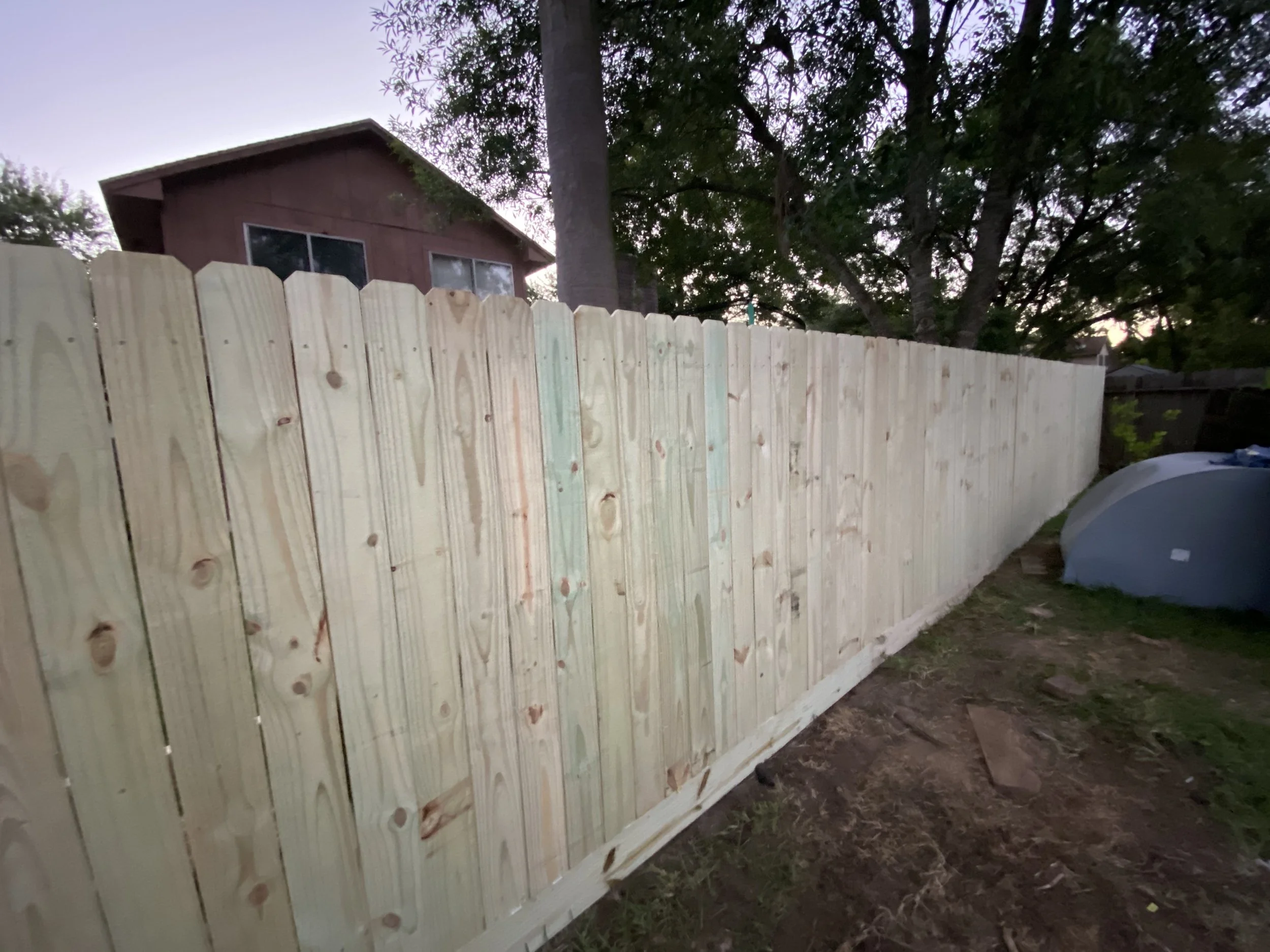 A wooden fence in front of a house with windows, trees in the background, and a car partially visible on the right side during dusk.