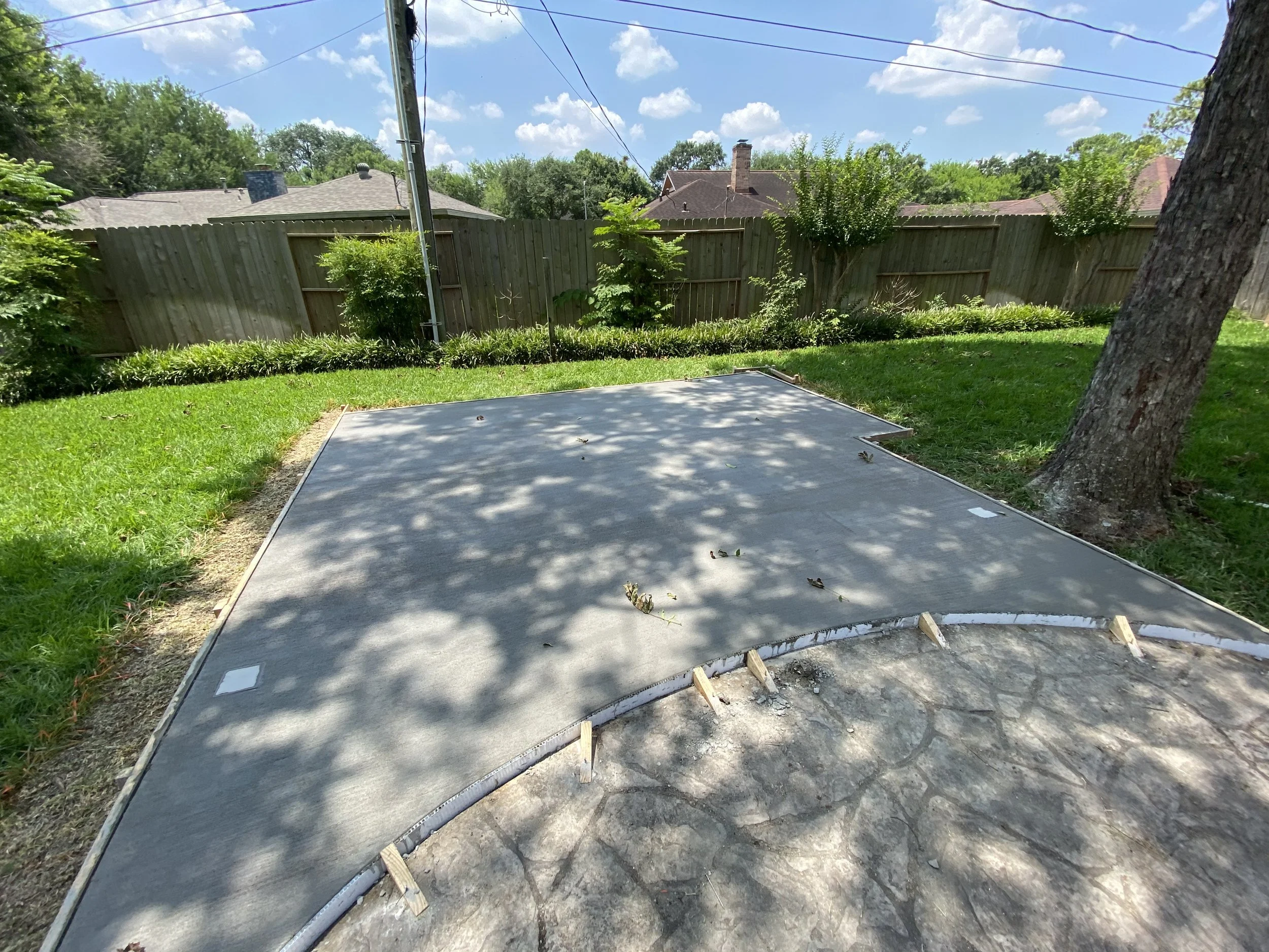 A backyard with a concrete slab foundation, surrounded by green grass, trees, a wooden fence, and suburban houses under a blue sky with clouds.