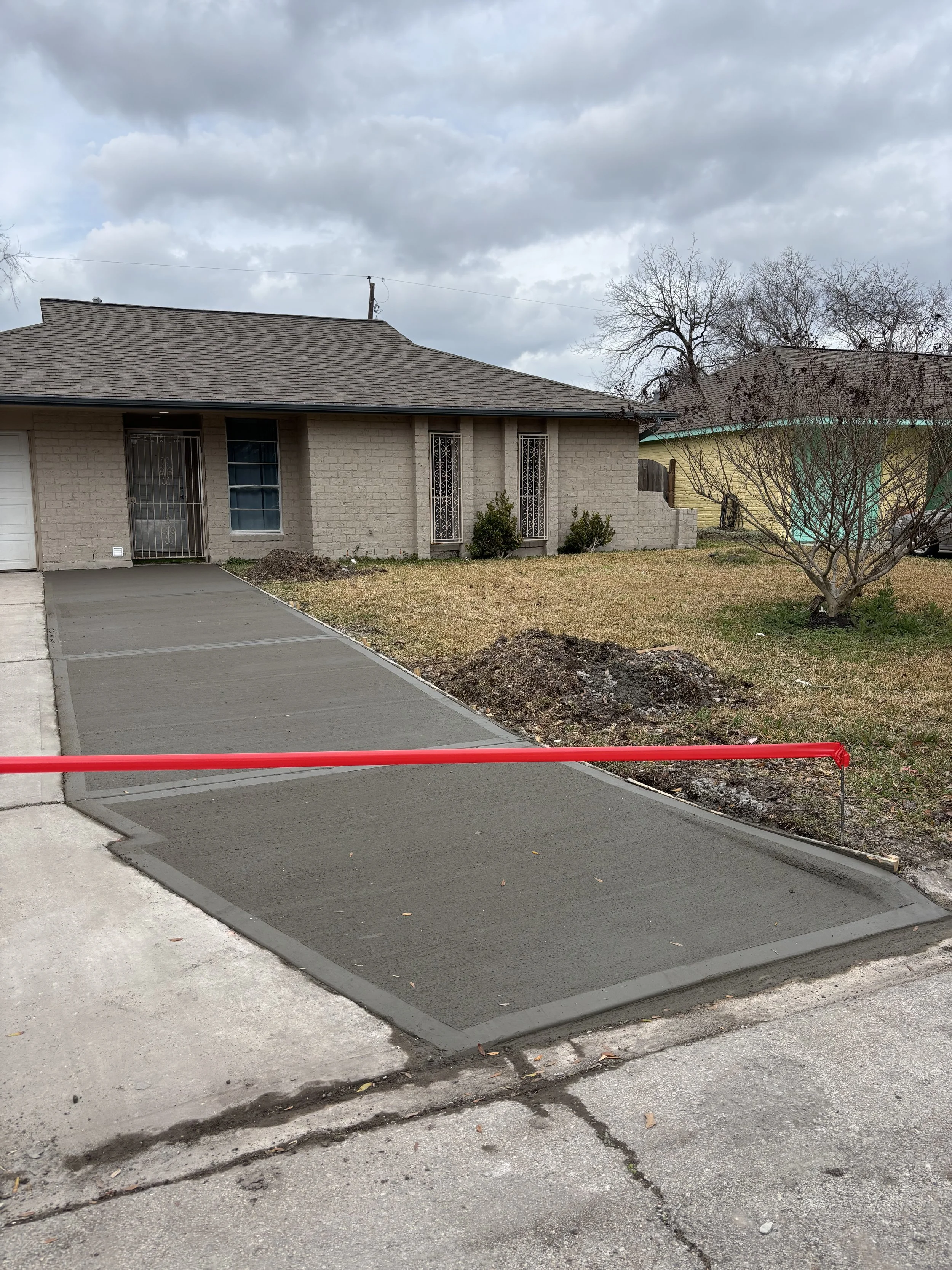 A freshly paved sidewalk with a red barrier at the edge in front of a residential house with a front yard and some bushes, under a cloudy sky.